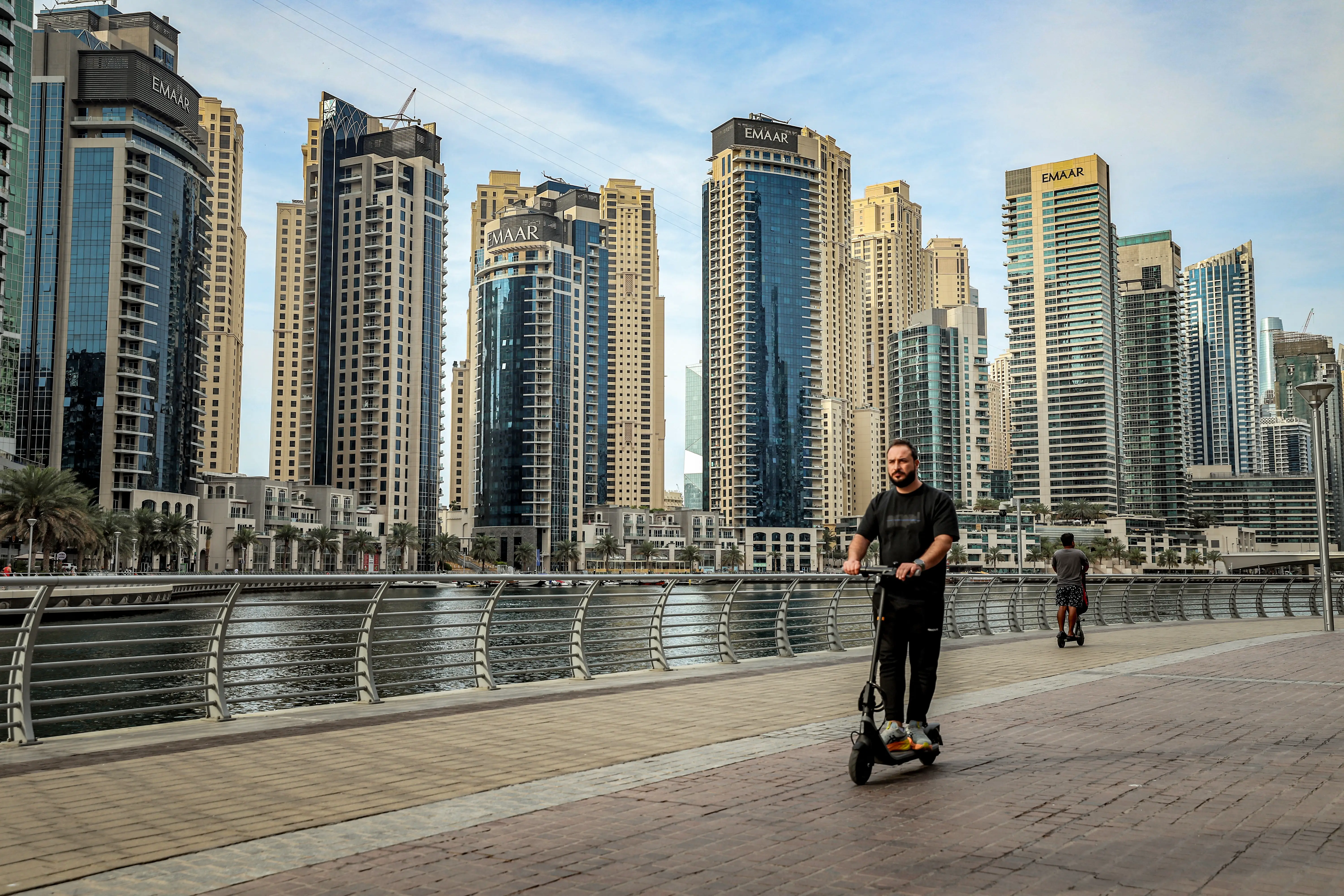 A man drives an electric scooter by the Dubai Marina against a backdrop of skyscrapers.