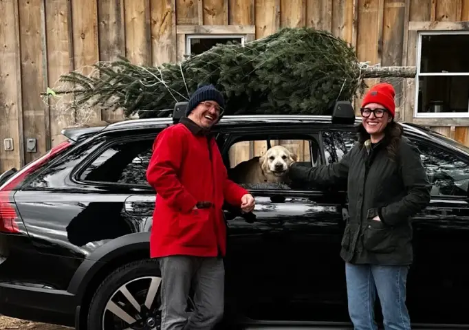 The writer, her husband, and their dog standing in front of a car and a Christmas tree.