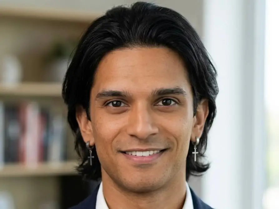 Smiling man with shoulder-length dark hair and cross earrings wears a navy blazer and white shirt in an indoor office setting.