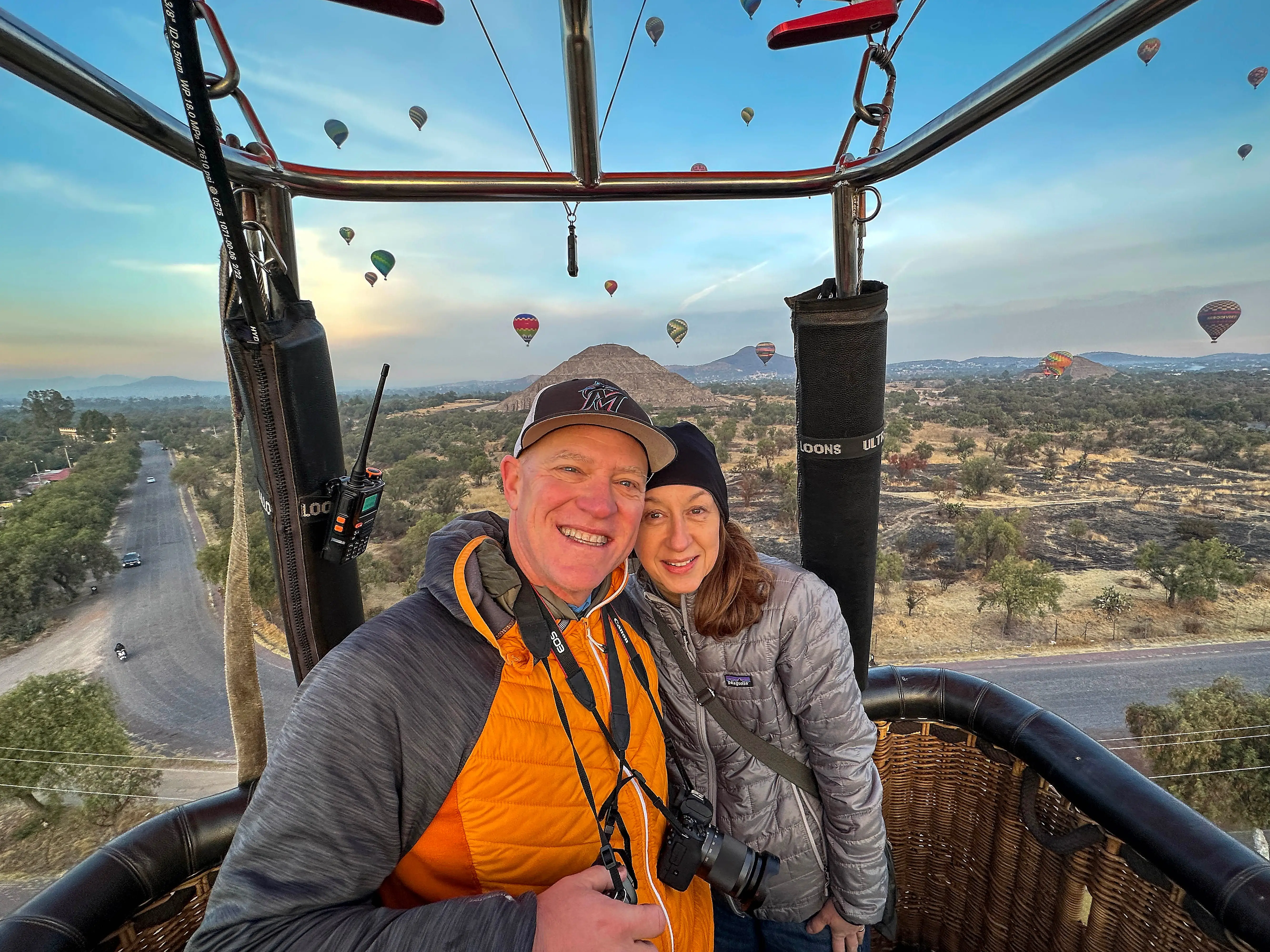 Thomas and his wife pose in a hot air balloon, with other balloons in the background.