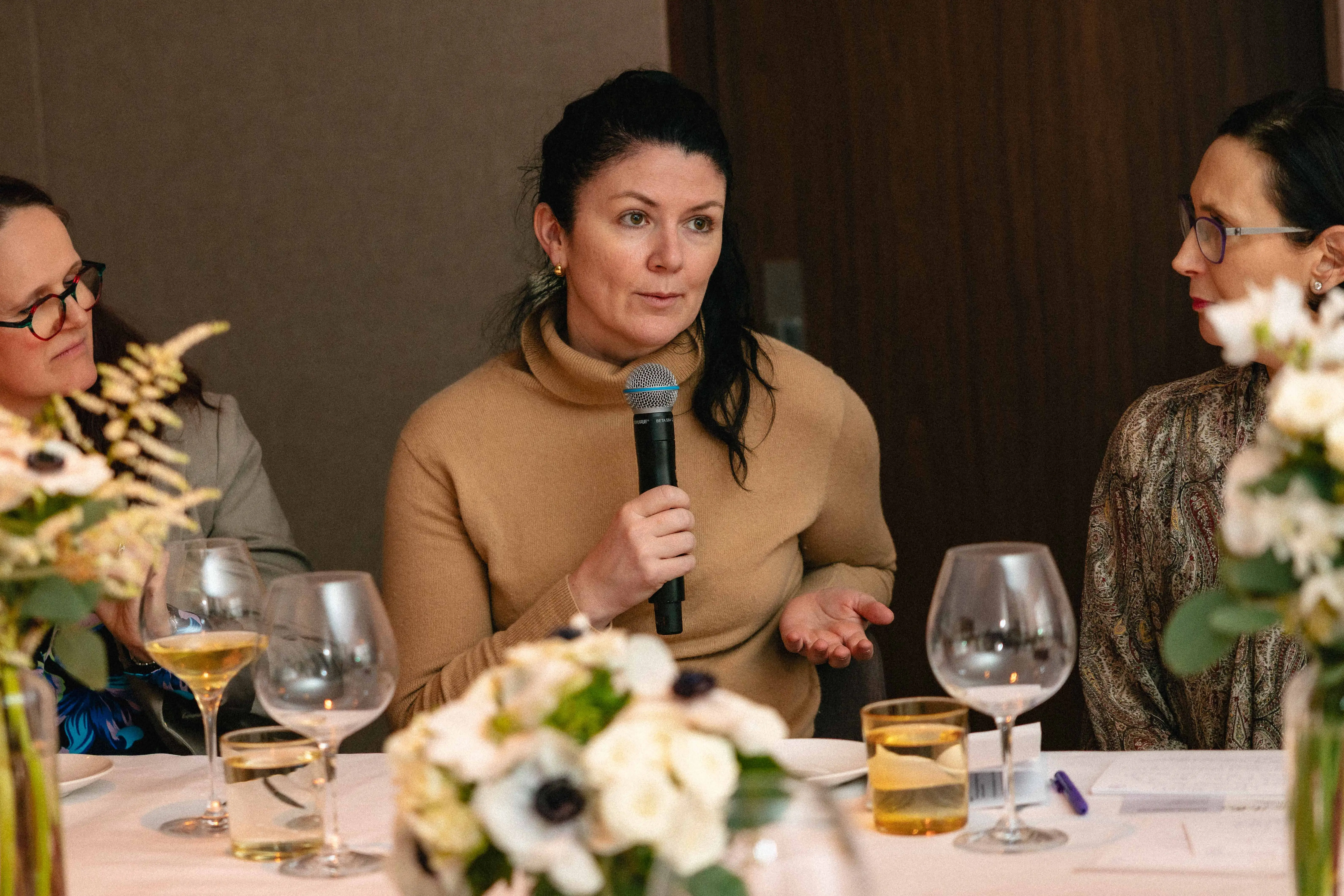 Woman in brown sweater at formal dinnertable speaking with microphone.