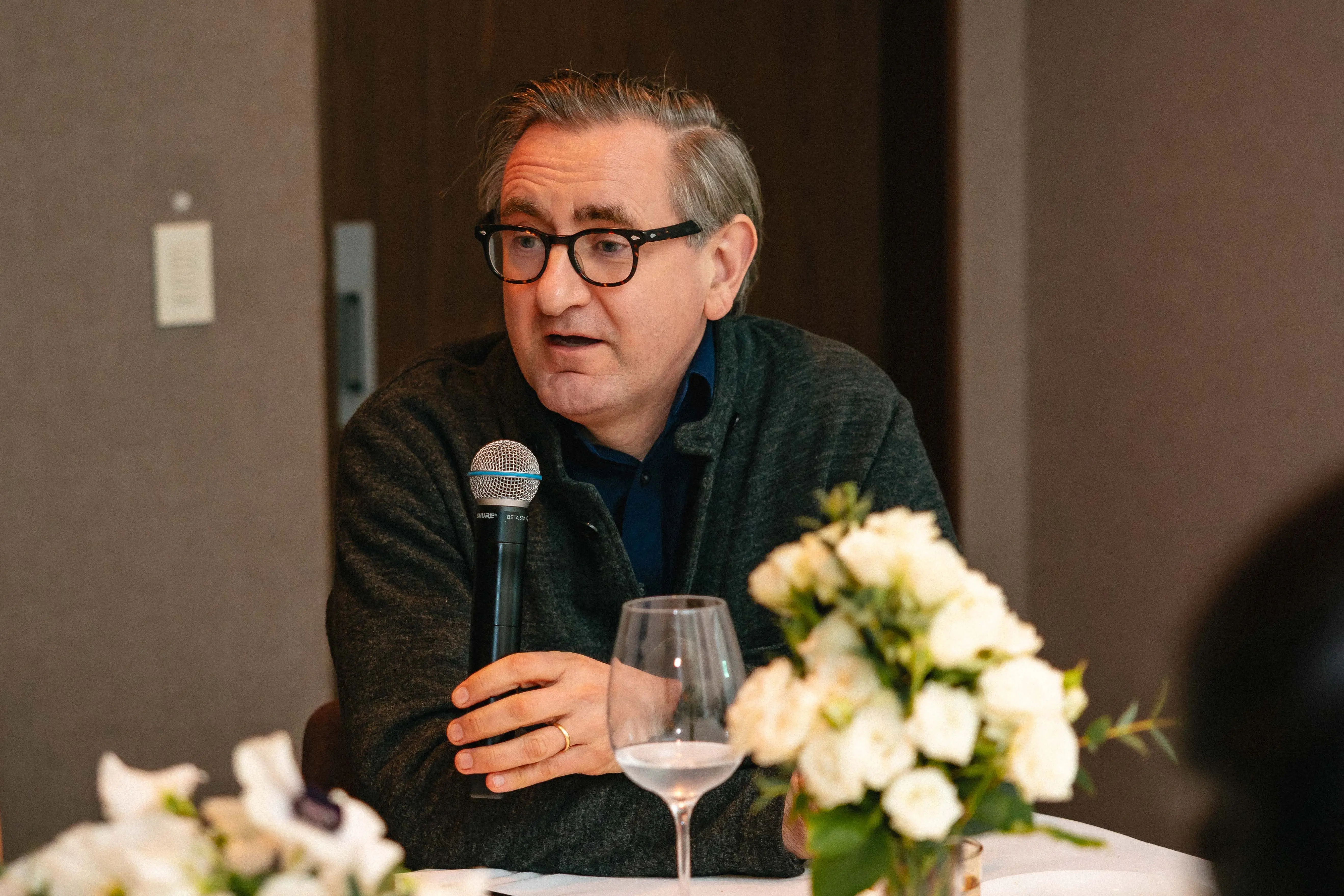 Older white man in glasses speaking into a microphone at a formal diner table.