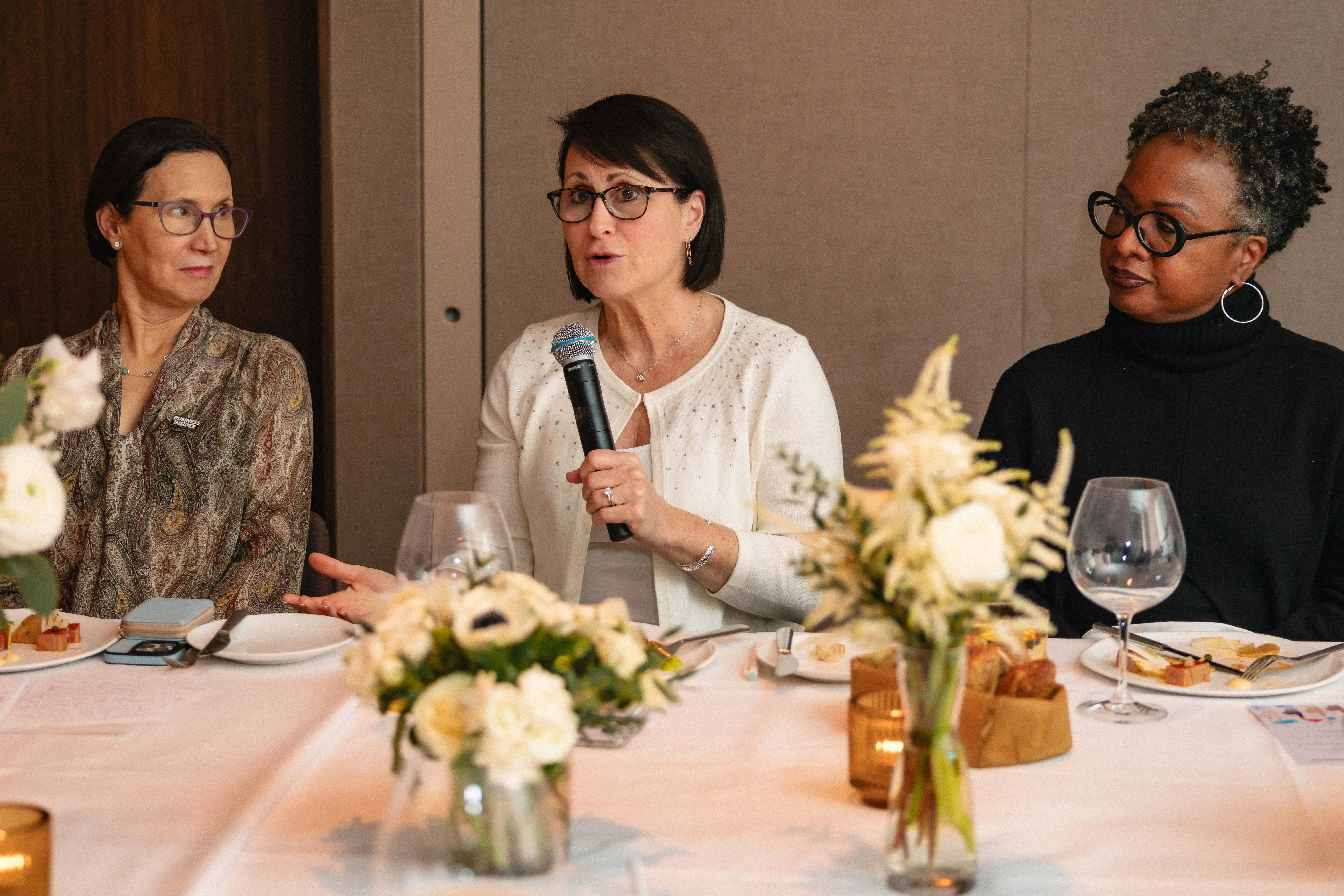Woman in white sweater with dark hair and glasses speaks into a microphone at a formal dinner table.