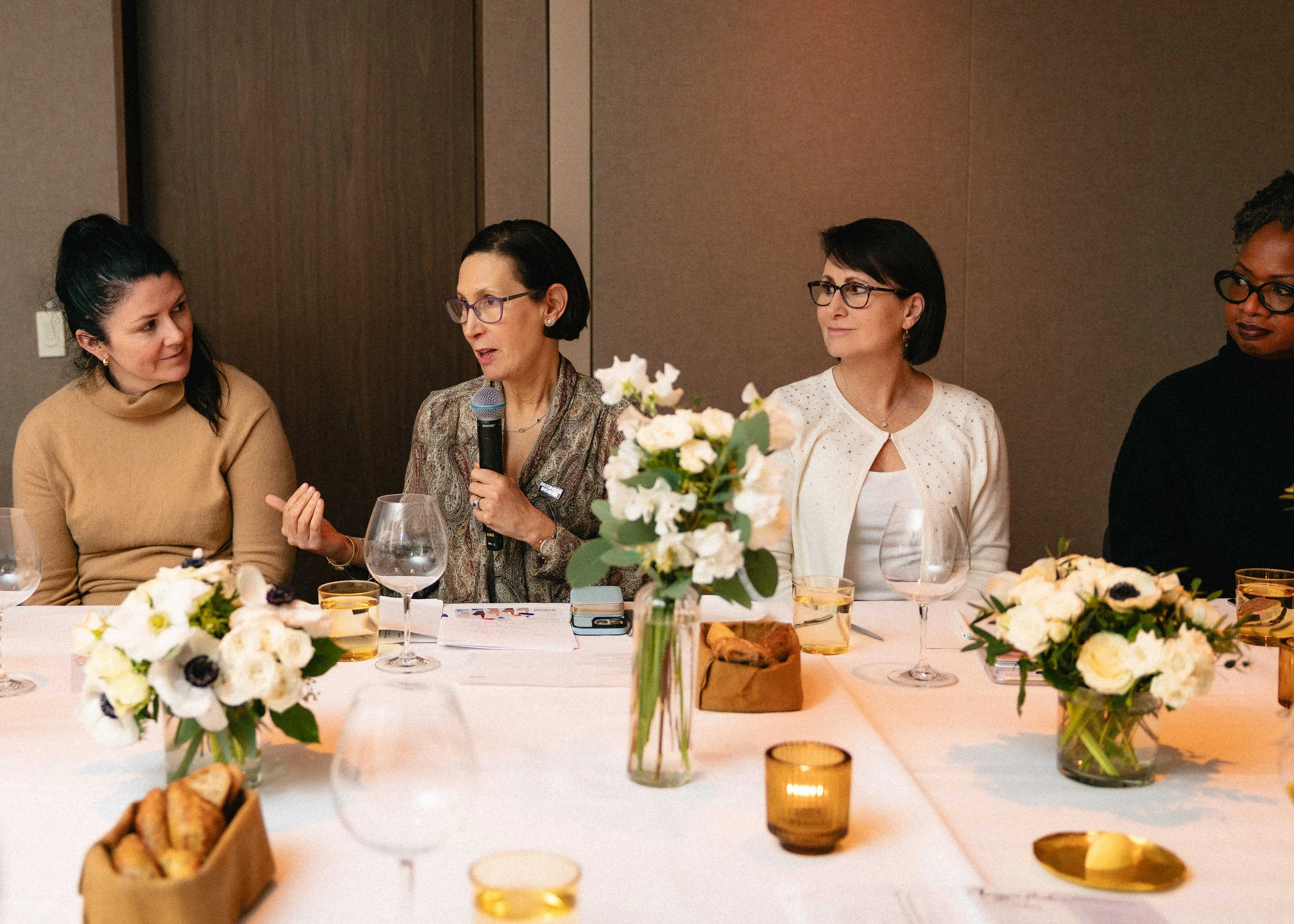 Three women at a long dinner formal dinner table listen to a fourth woman who is holding a microphone and speaking.