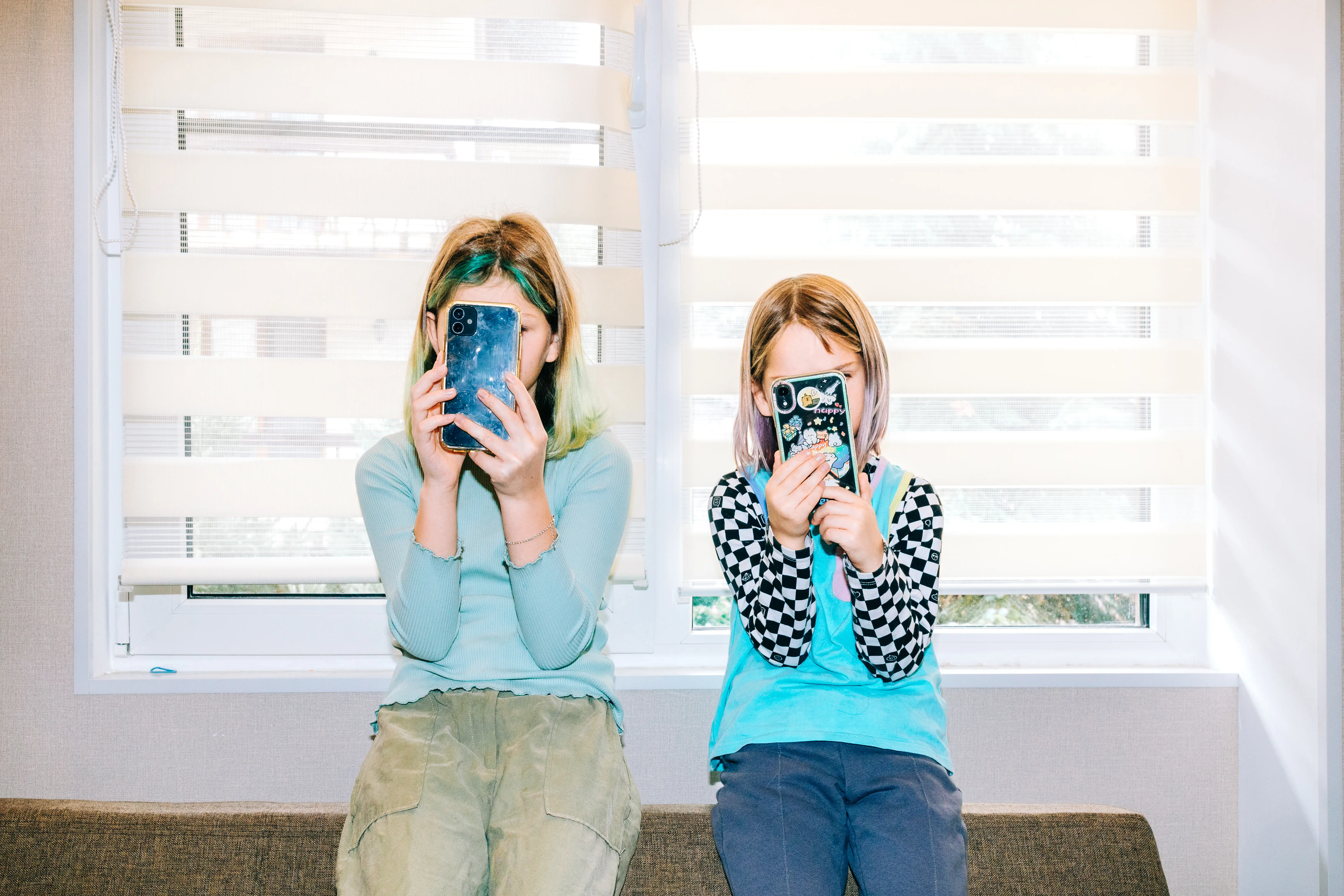 Two kids sit on a bench in front of a windo with smartphones obscuring their faces.