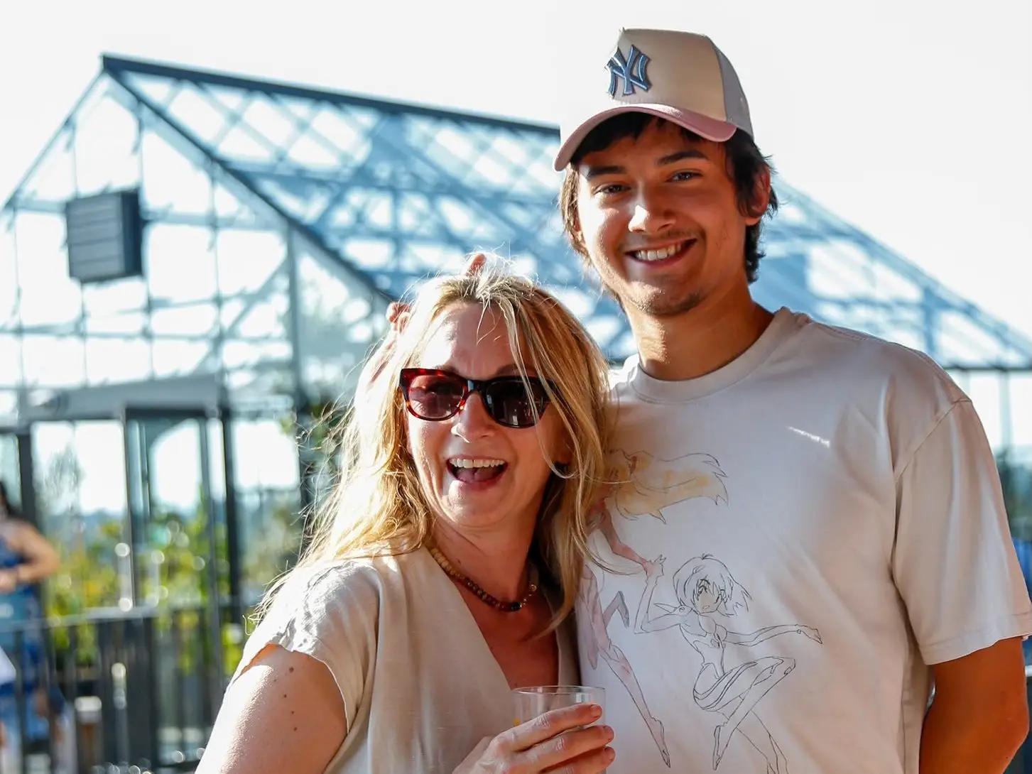 A woman poses next to her son, they smile in front of a greenhouse.