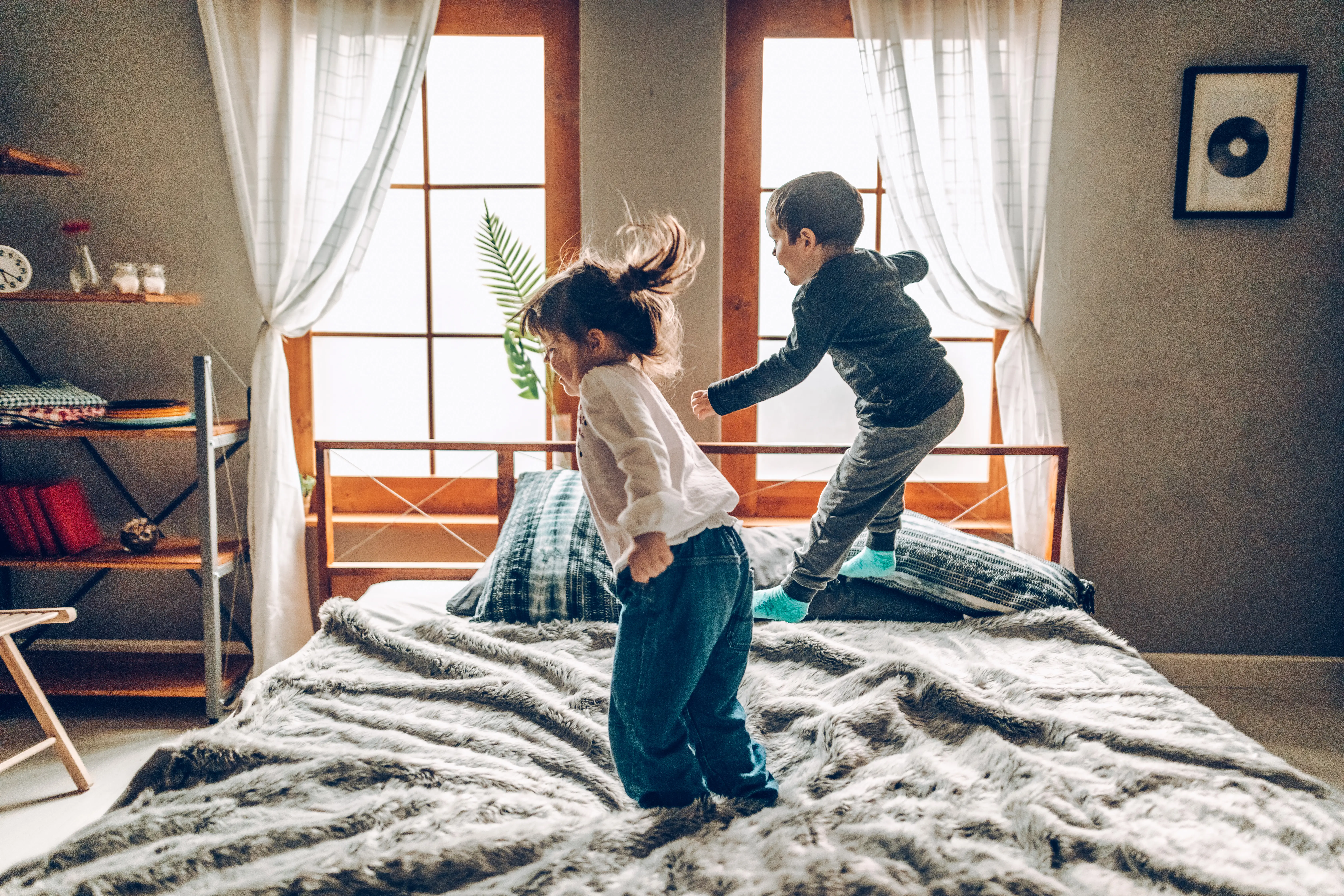 Two kids jumping on a bed with windows in the background.