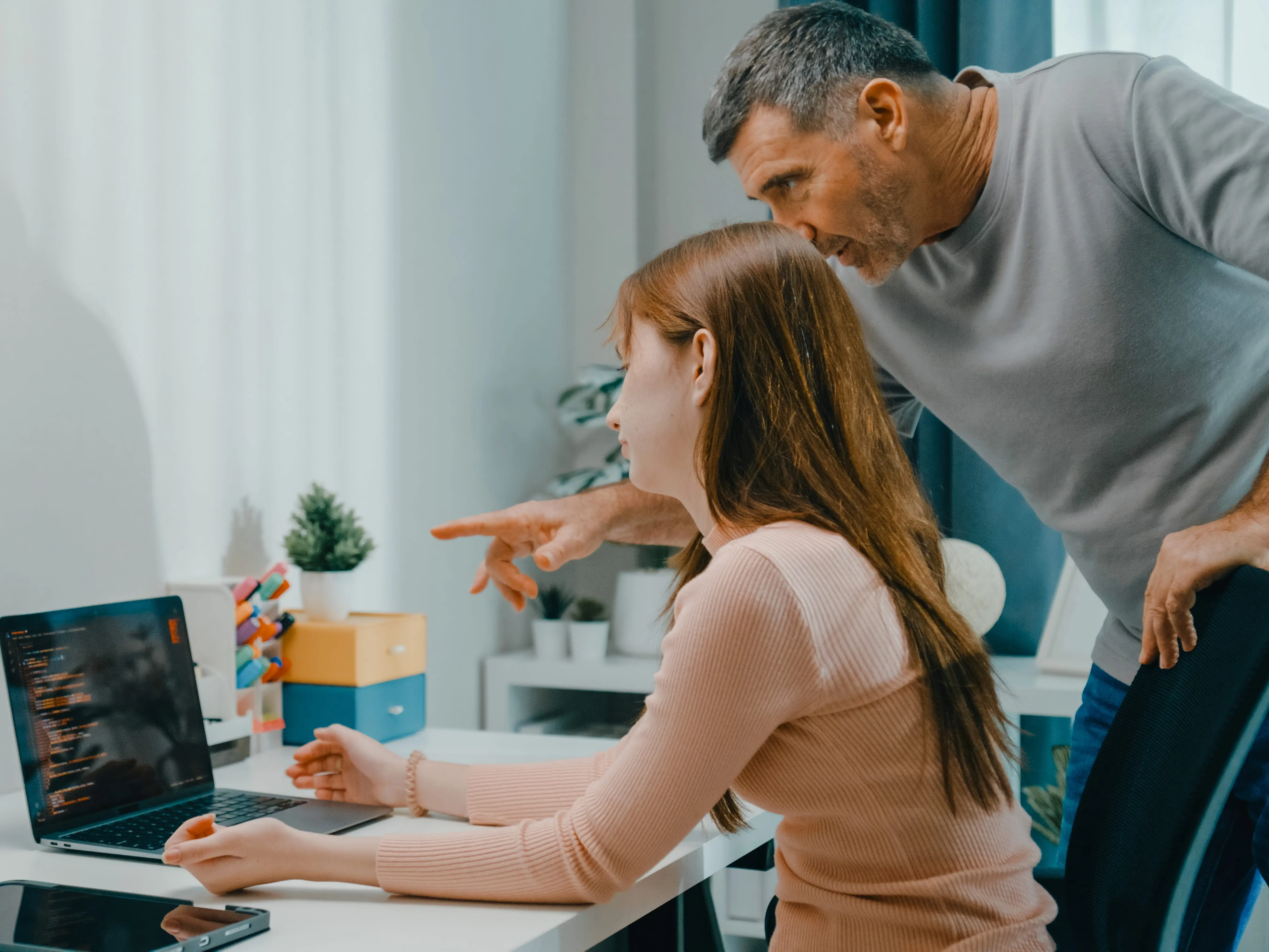 A father leans over a teens shoulder as she works on a laptop.