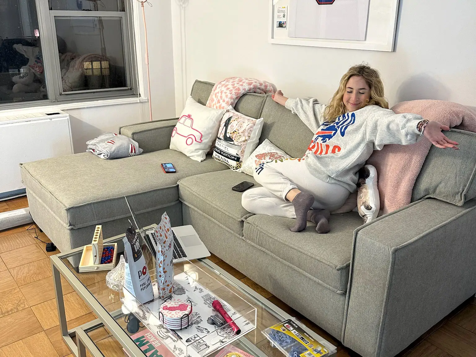 Carrie, with her arms stretched out to the sides, sits on a couch in her apartment.
