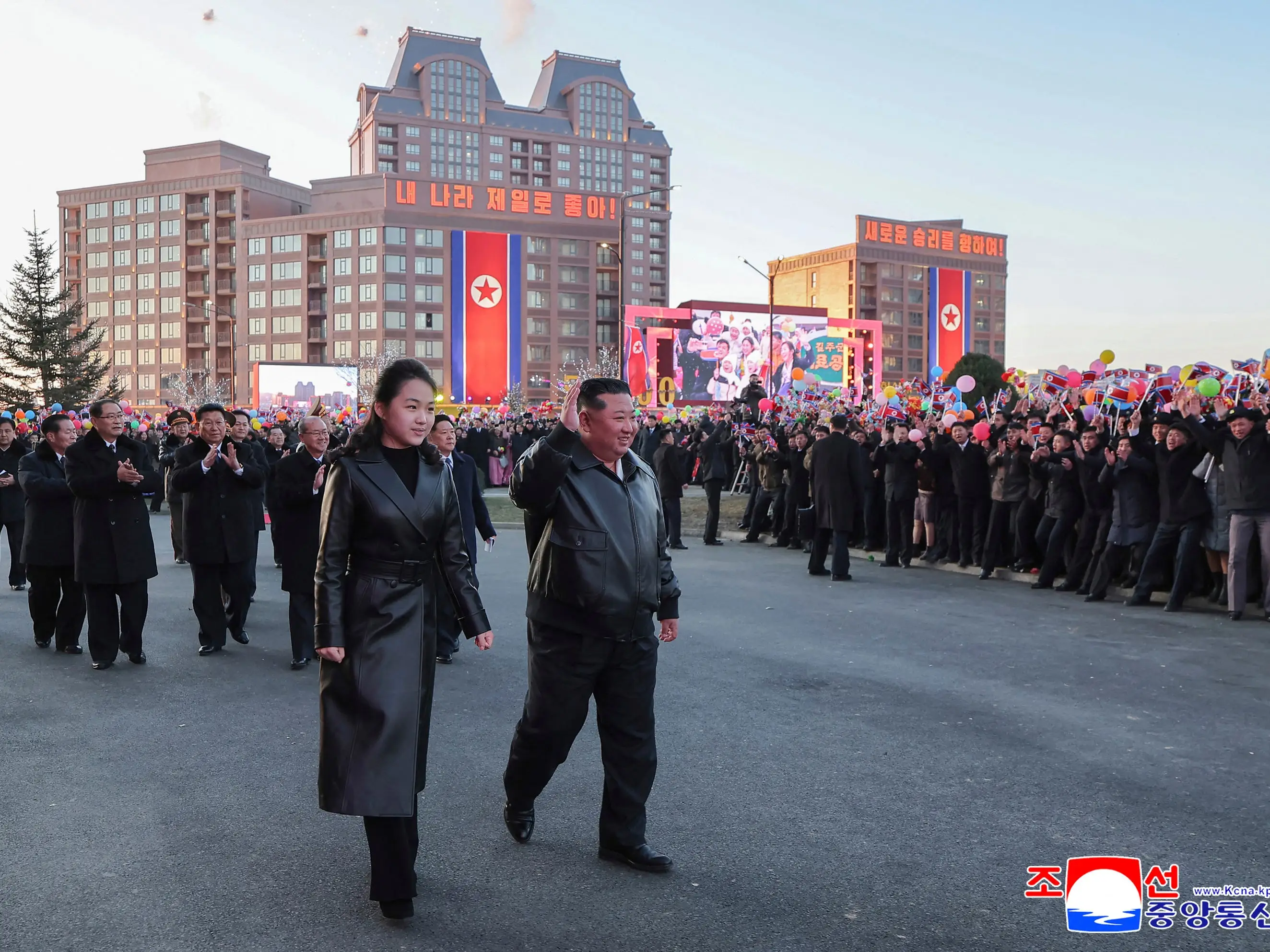 Kim Jong Un and Kim Ju Ae walking amongst a large crowd with buildings in the background.