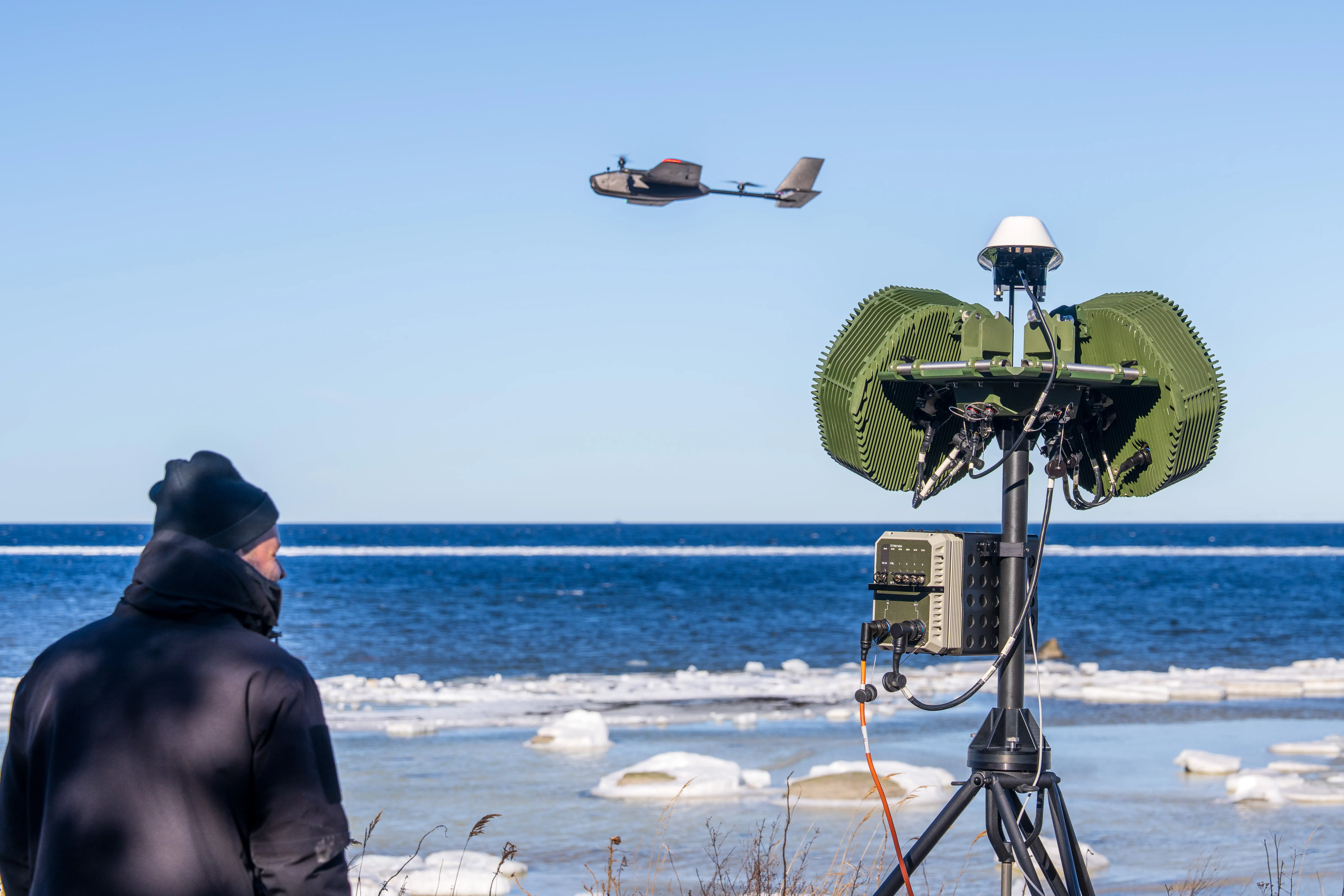 A man stands next to a tall radar. An uncrewed system flies in the sky past him. The beach is icy.