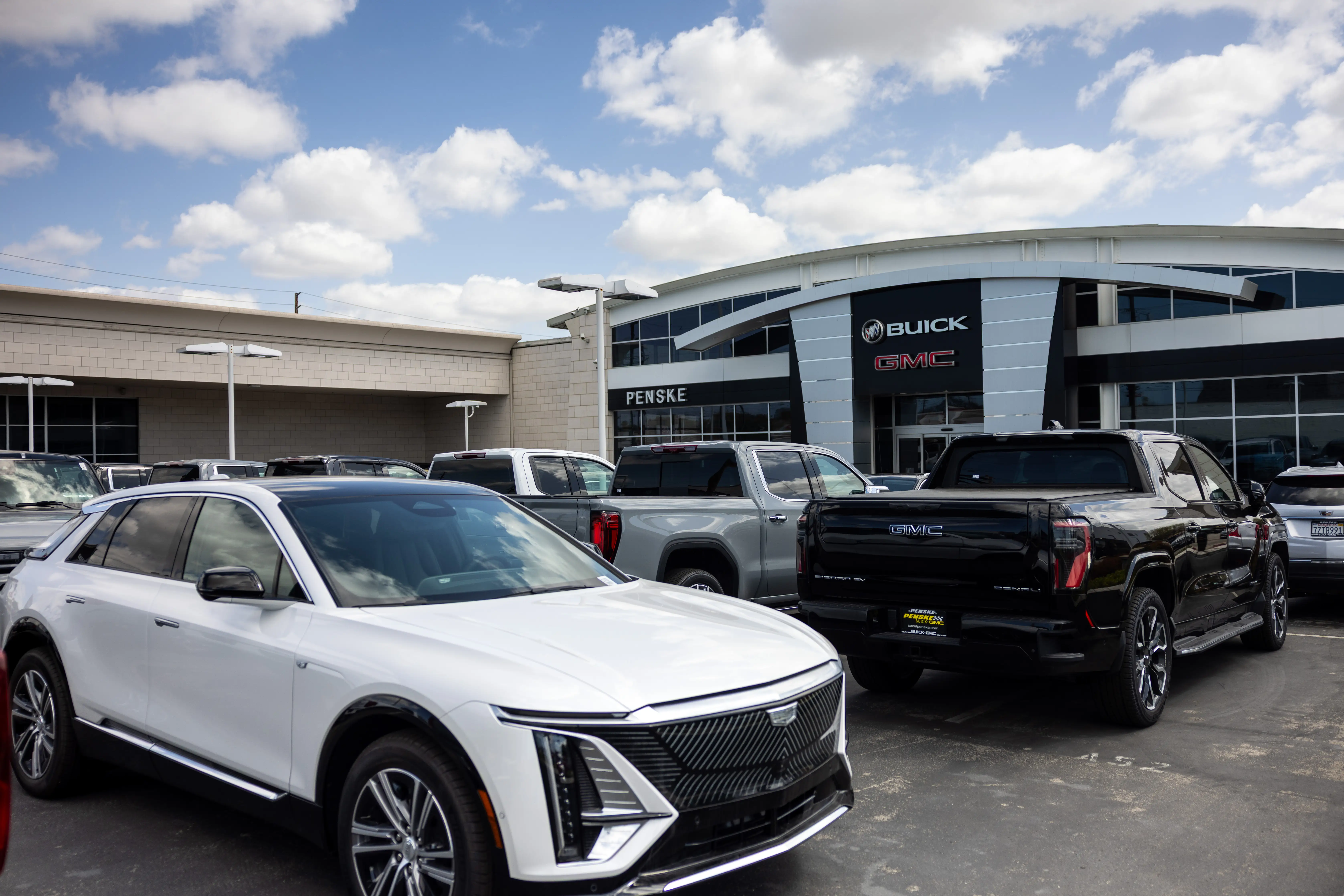 A white Cadillac Lyric EV sits behind a row of GMC Sierra EVs on a GM dealership lot.