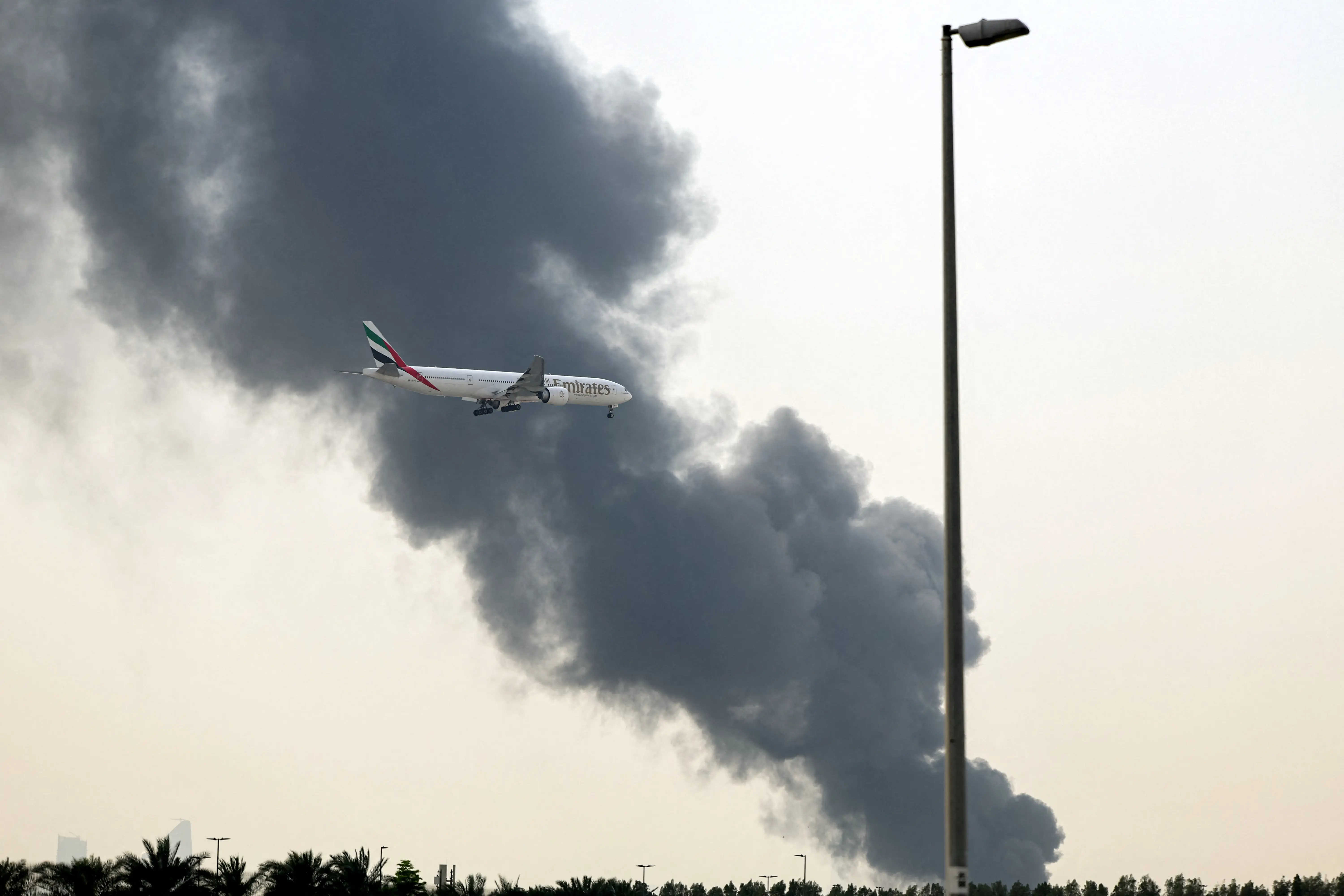 An Emirates Boeing 777 aircraft prepares for landing as a smoke plume rises from an ongoing fire near Dubai International Airport in Dubai on March 16, 2026.