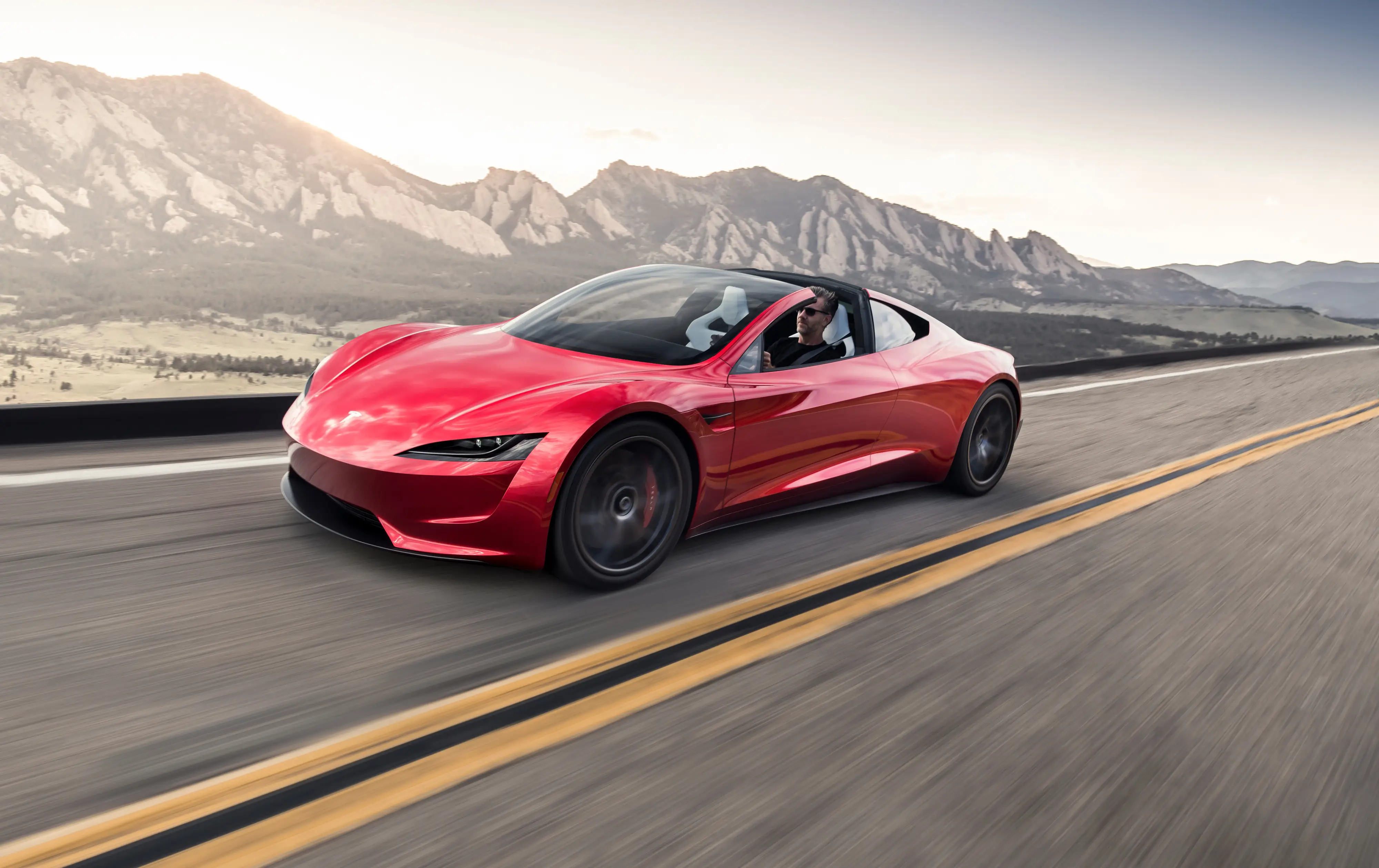 A red Tesla Roadster drives down a two-lane road next to a desert mountain range.