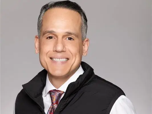 A man in a white shirt and tie smiles while wearing a black Webull-branded vest against a gray background.