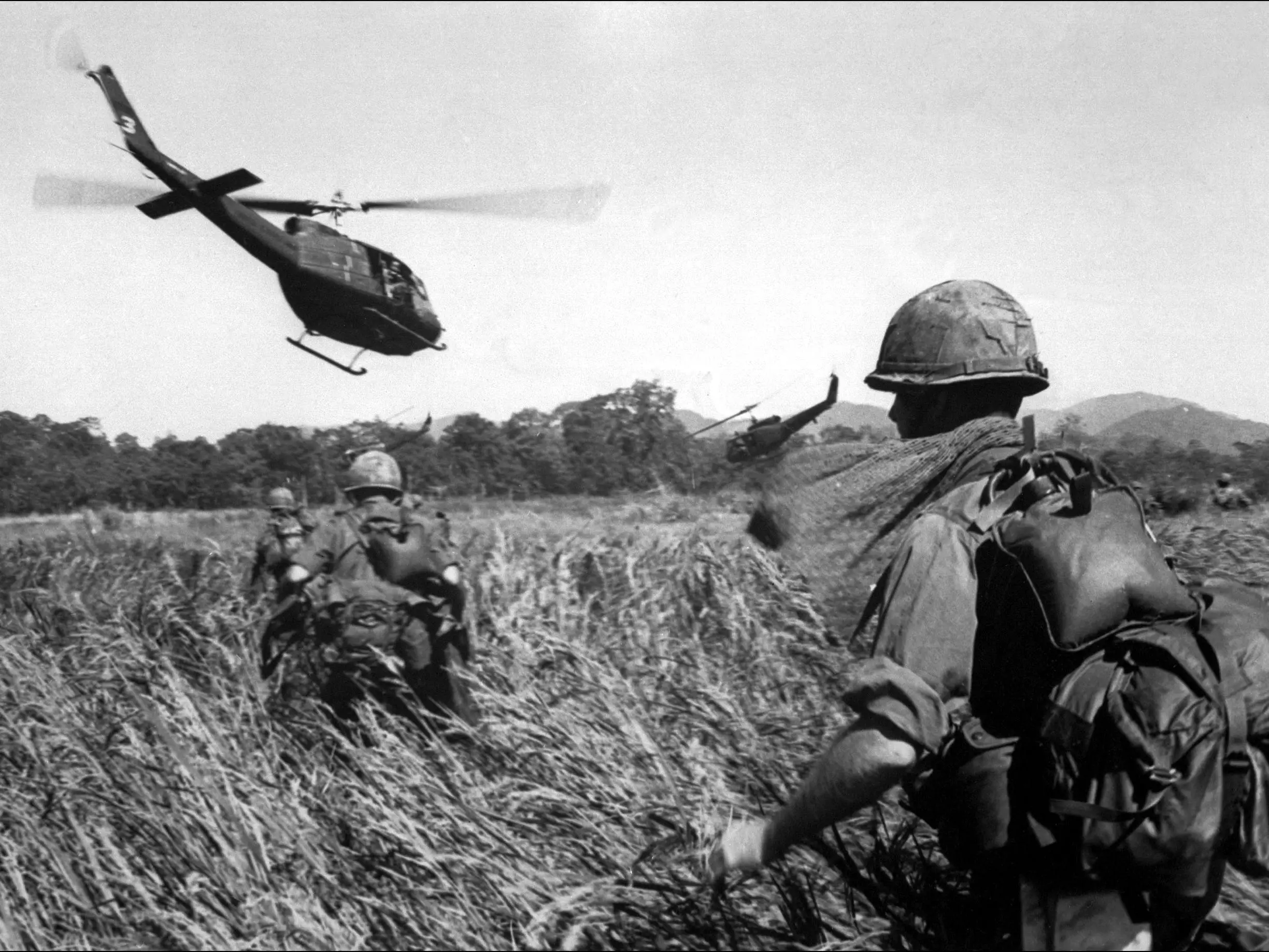 American soldiers walking in tall grass while a helicopter flies overhead.