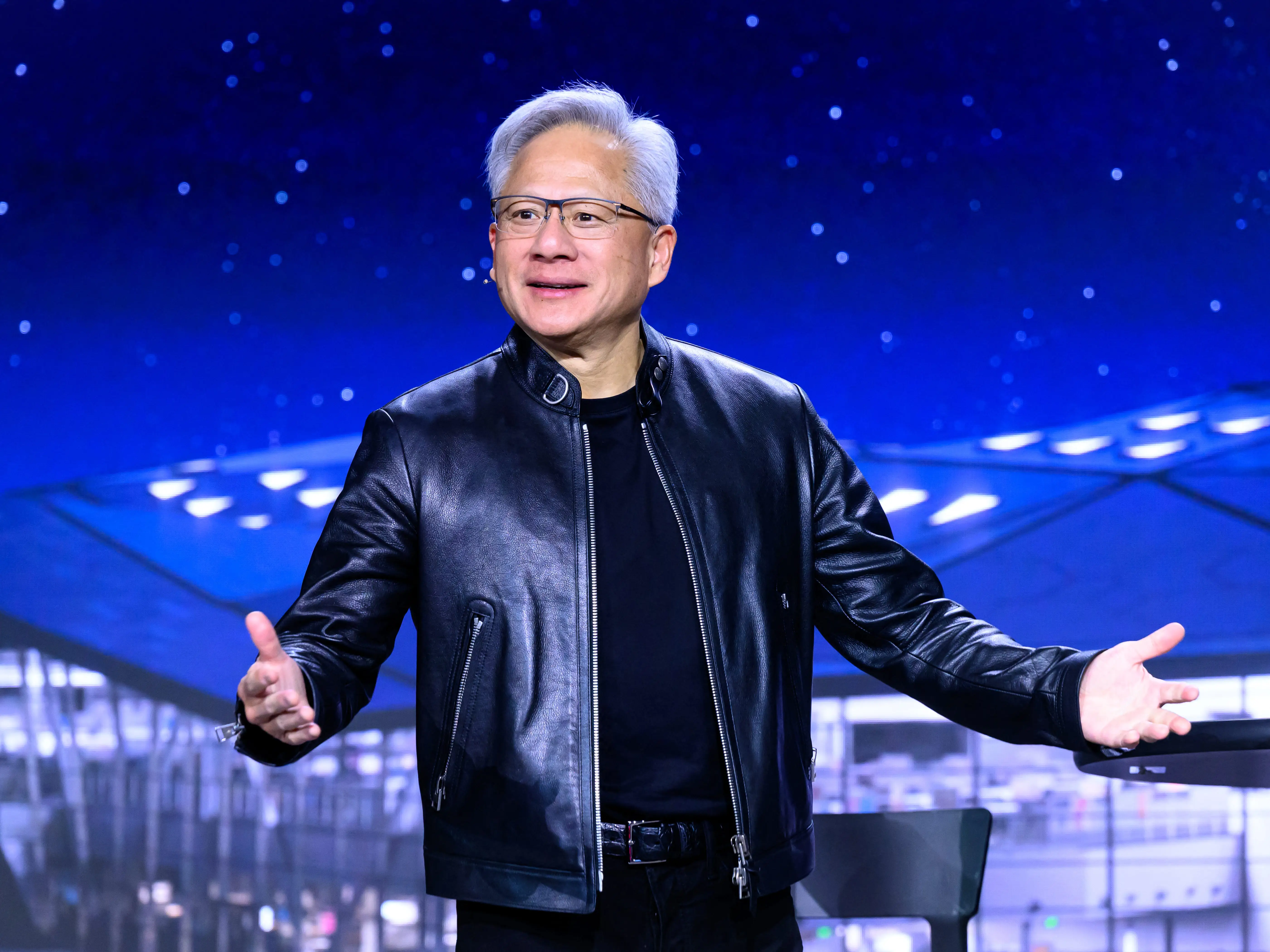 Jensen Huang onstage at GTC, wearing a black leather jacket standing in front of a blue background.