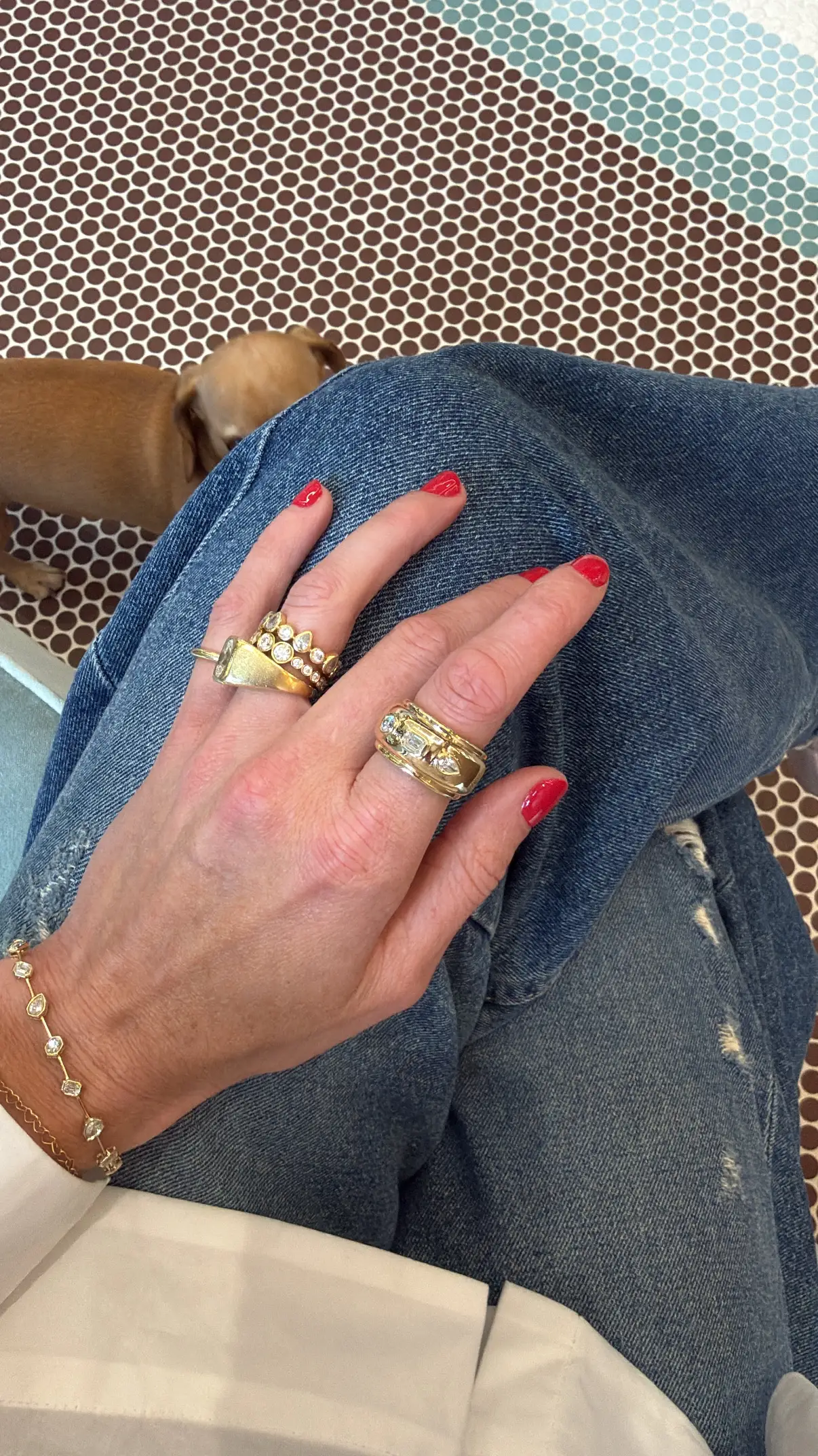 Hand with red manicured nails wearing four gold pieces of jewelry, including an Oura Ring jacket, as well as a diamond encrusted bracelet.