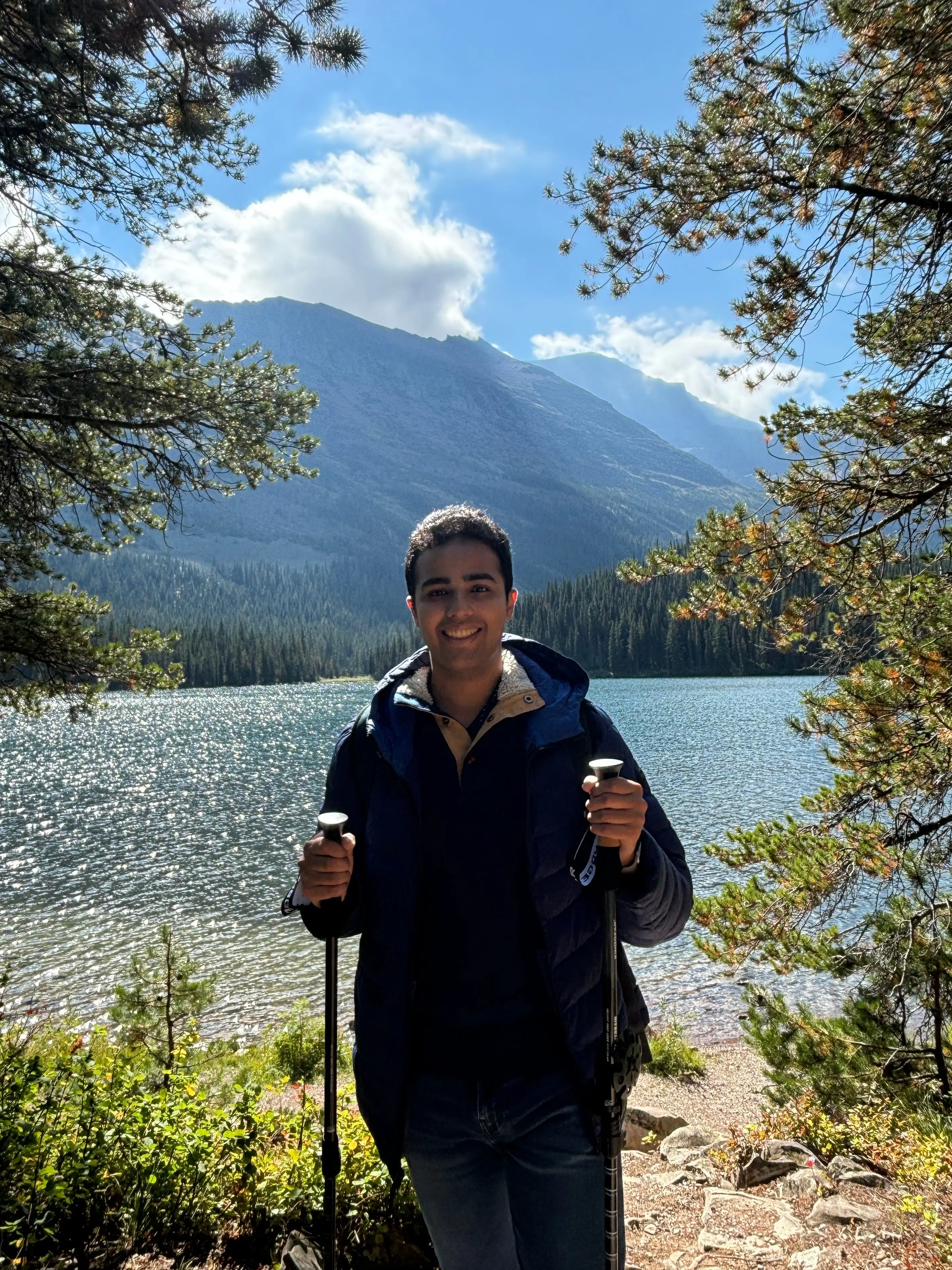 Kanav Bhatnagar is holding two walking poles in front of a view of an open body of water and a mountain.