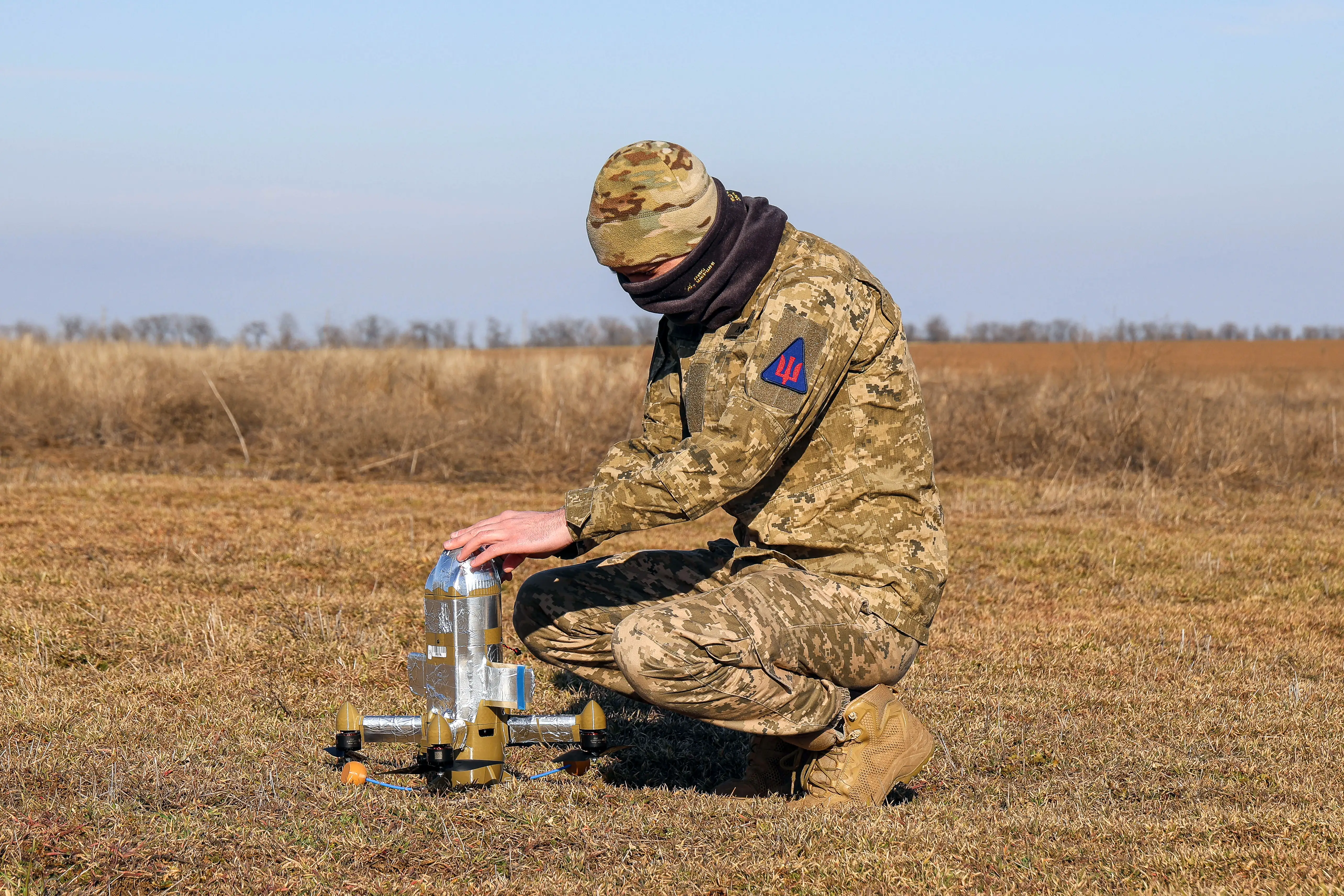 A man in camouflage gear squats in a yellow field holding a silver drone