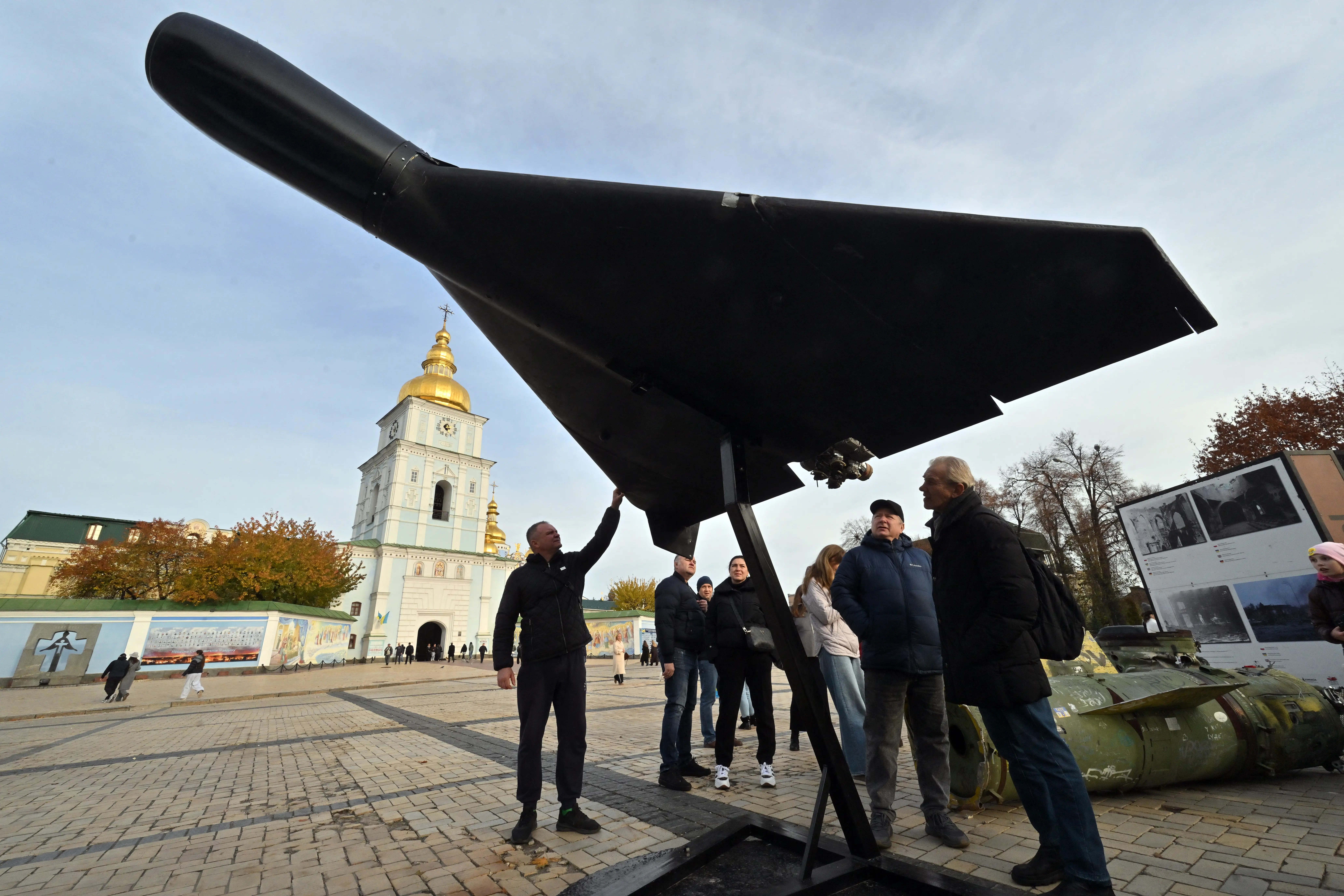 People gesture up to a large black drone on display on a street with a gold-topped church in the background