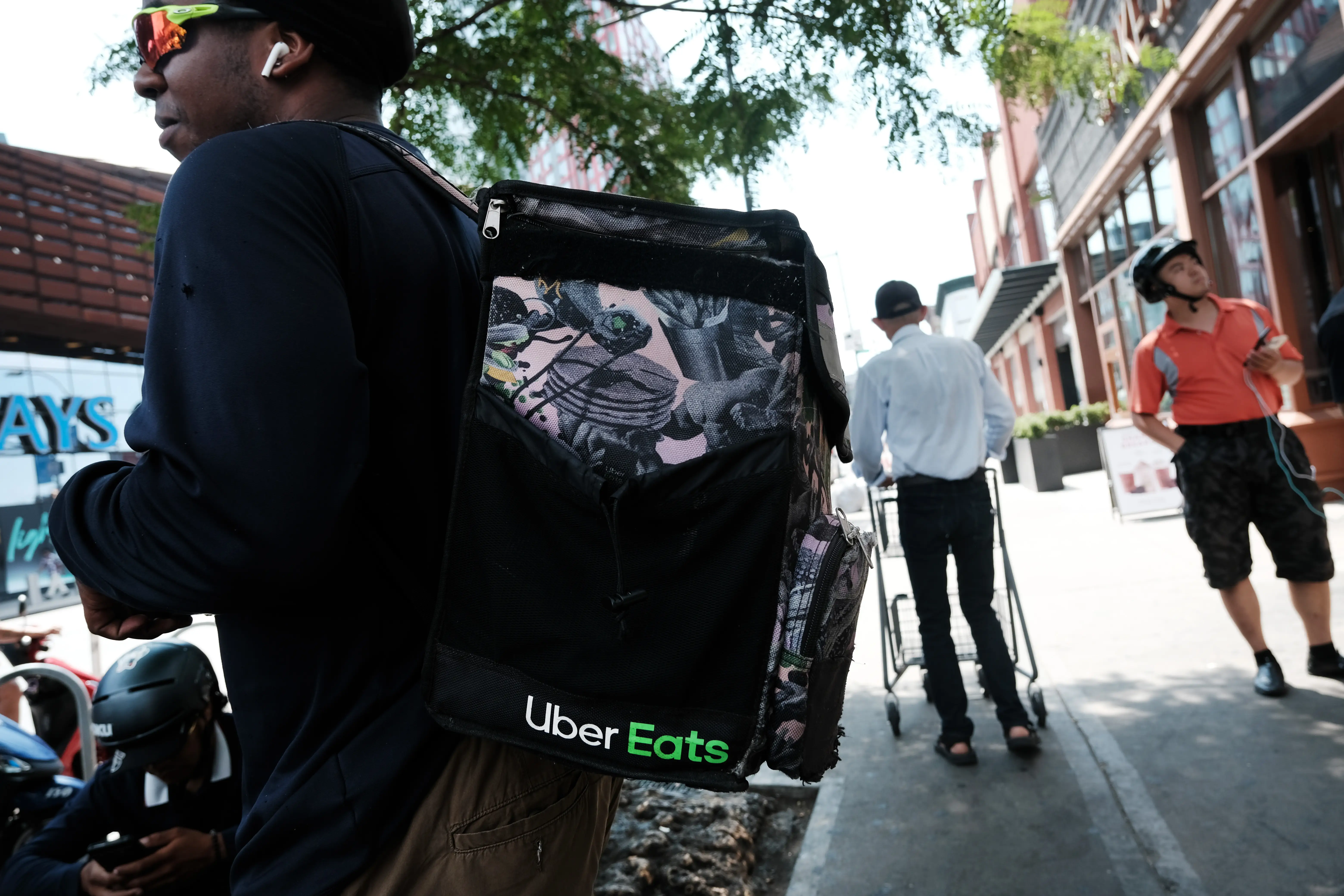 An Uber Eats delivery worker stands in Brooklyn, New York wearing sunglasses and a black Uber Eats-branded backpack as pedestrians walk behind him on the street.