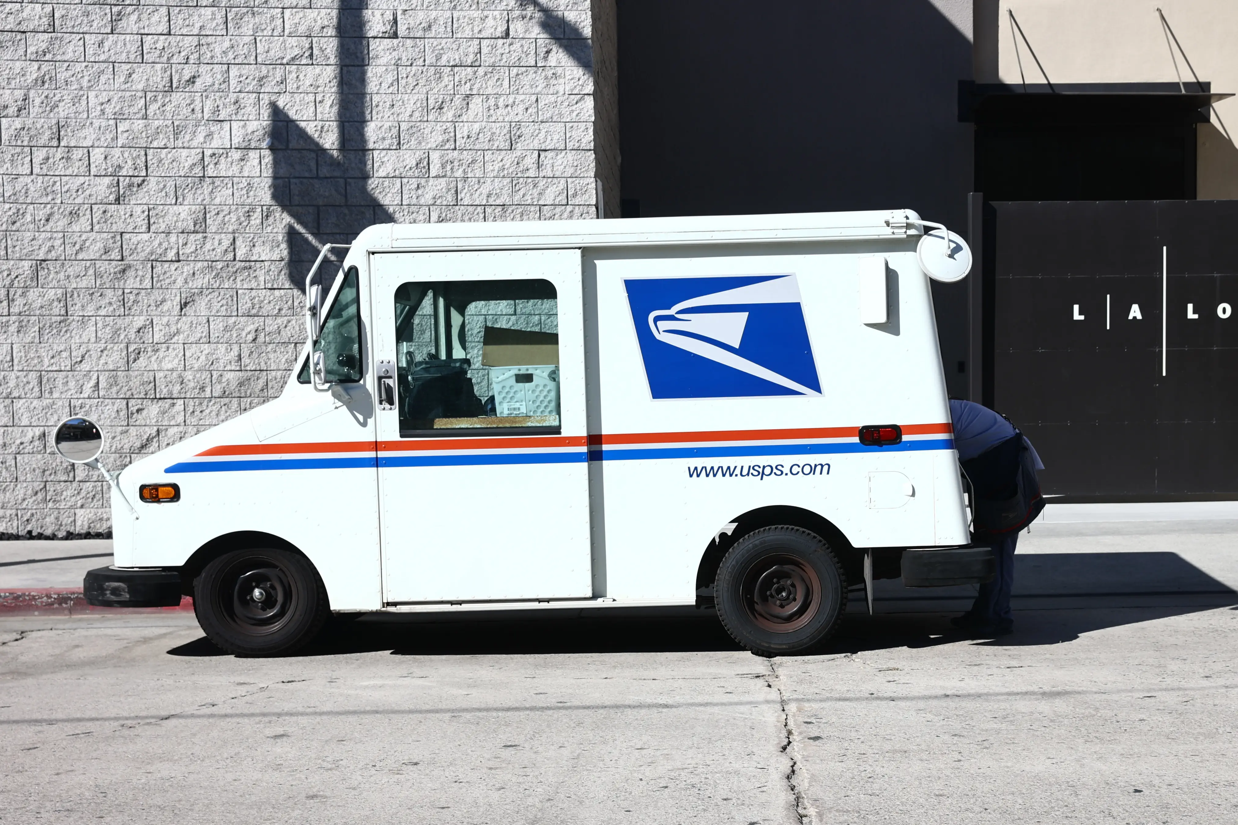 A mailman reaches into the back of a small, white USPS truck.