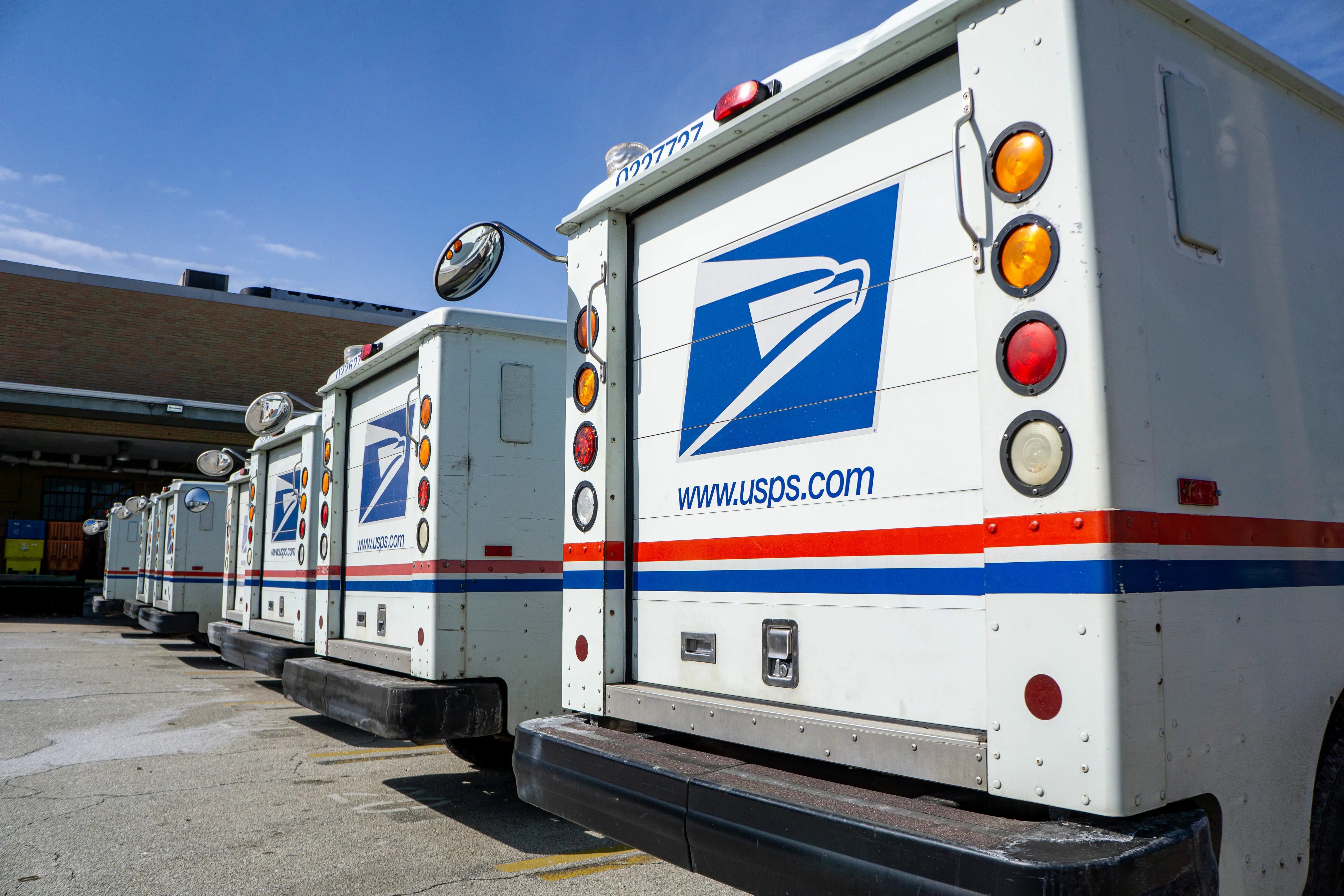 A line of USPS trucks from the back outside of a brick building.