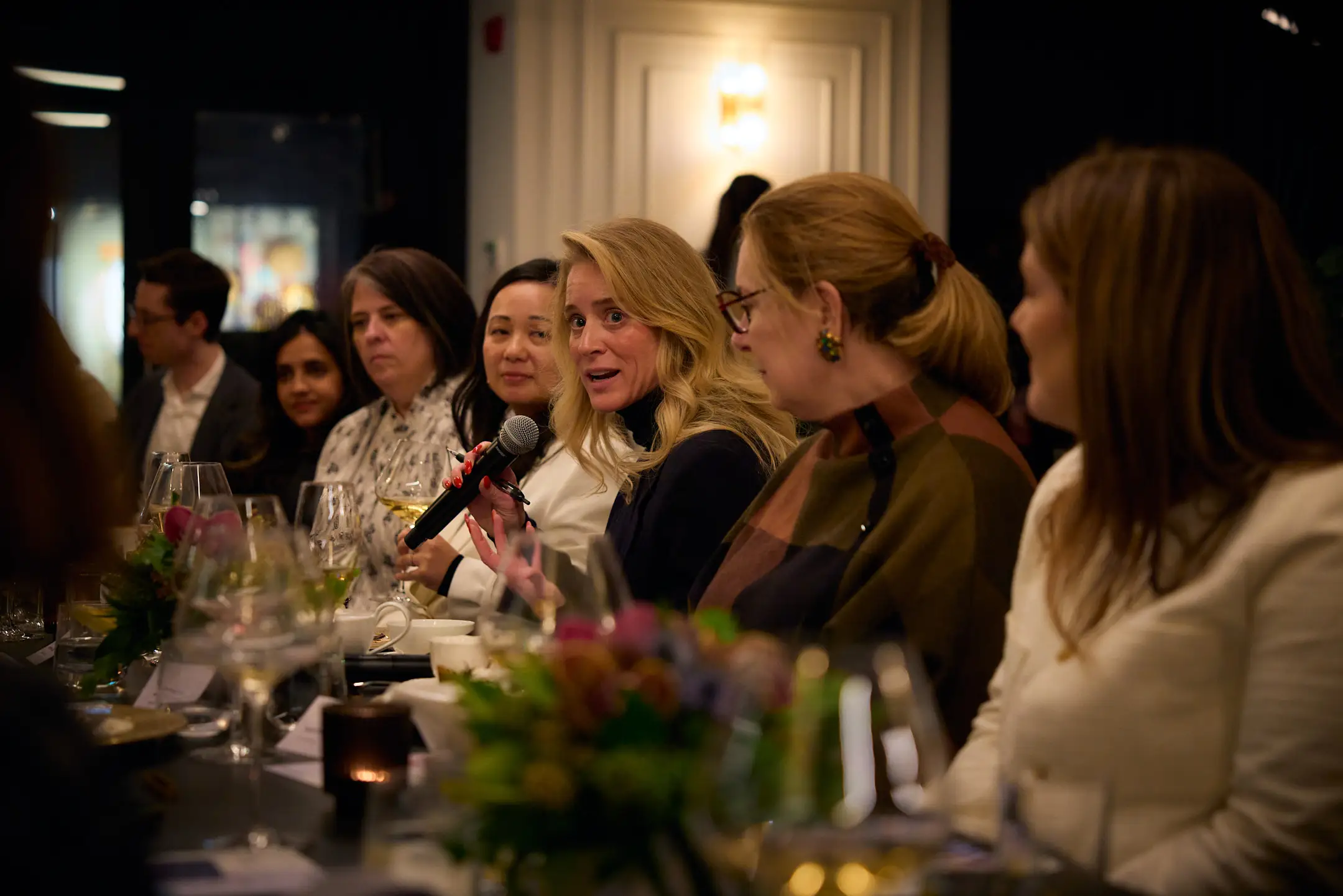 Blond woman holding a microphone and sitting at a long table speaks to group.