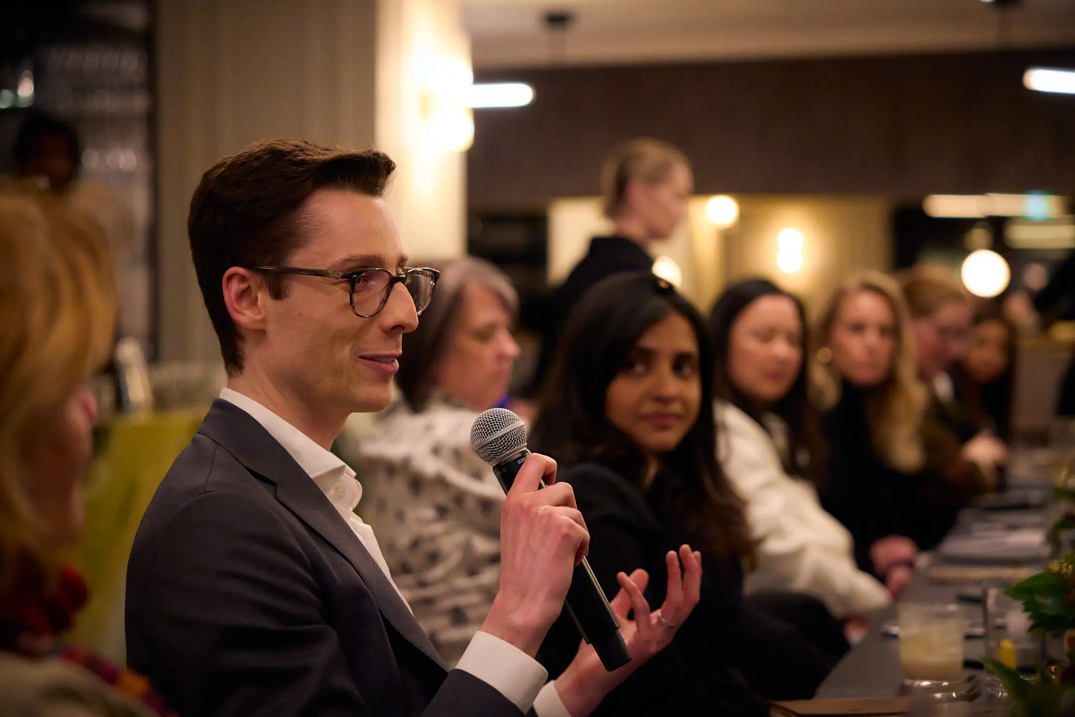 Man in glasses holding a microphone and seated at a long table speaks to a group.