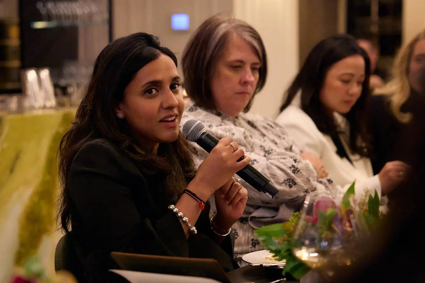 Dark-haired woman holding a microphone and sitting at a long table speaks to a group.