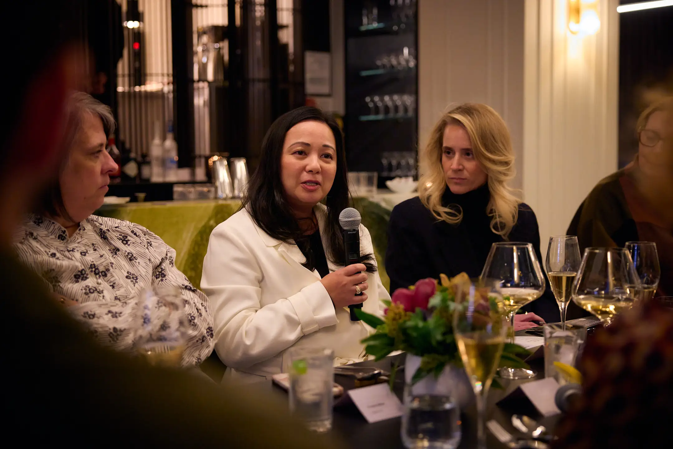 Woman in white blazer holding a microphone and sitting at a table speaks to a group.