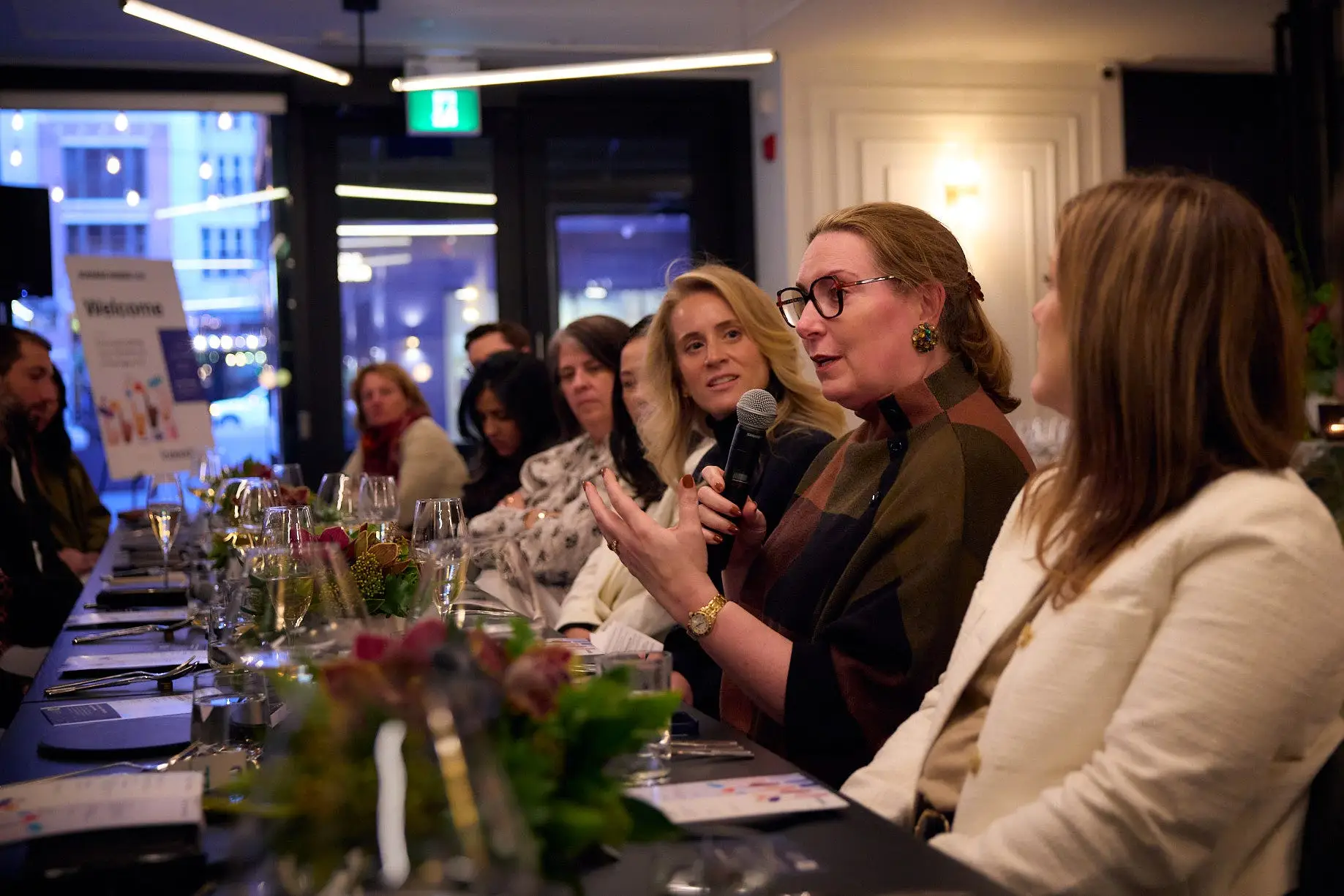Woman in glasses holding microphone and sitting at a long table speaks to a group.