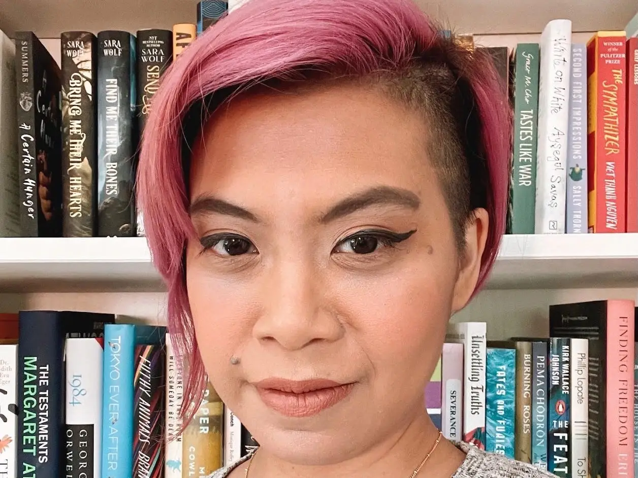 A person with short pink hair looks at the camera in front of a bookshelf filled with novels.