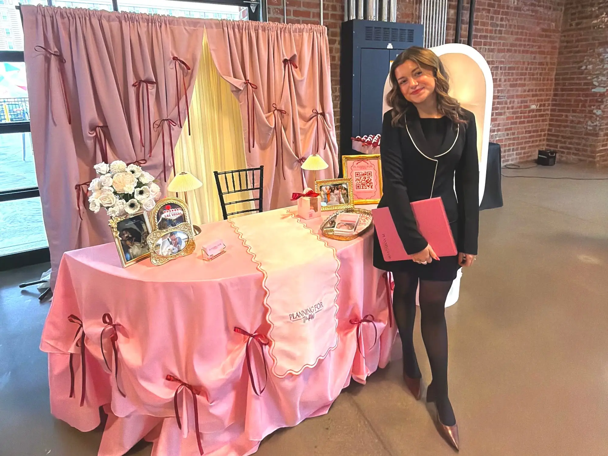 A woman in a black outfit stands in front of a table covered in pink drapery.