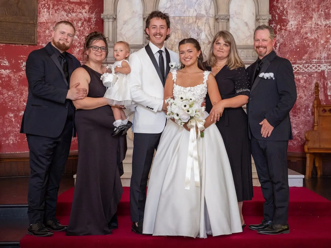 A bride and groom stand in a church with red decor next to their family members.