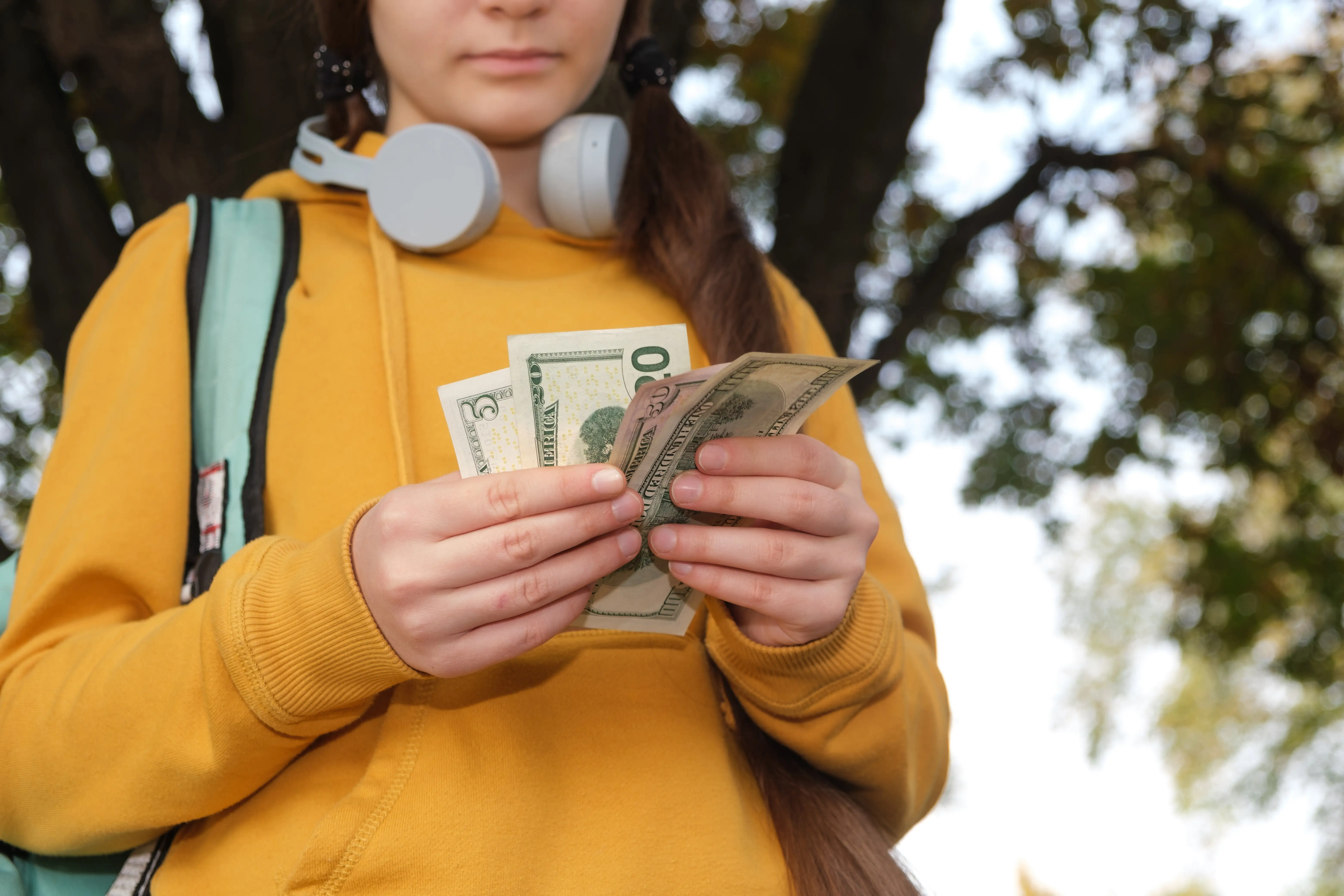 A teen wearing a yellow hoodie, headphones, and a backpack counts cash.