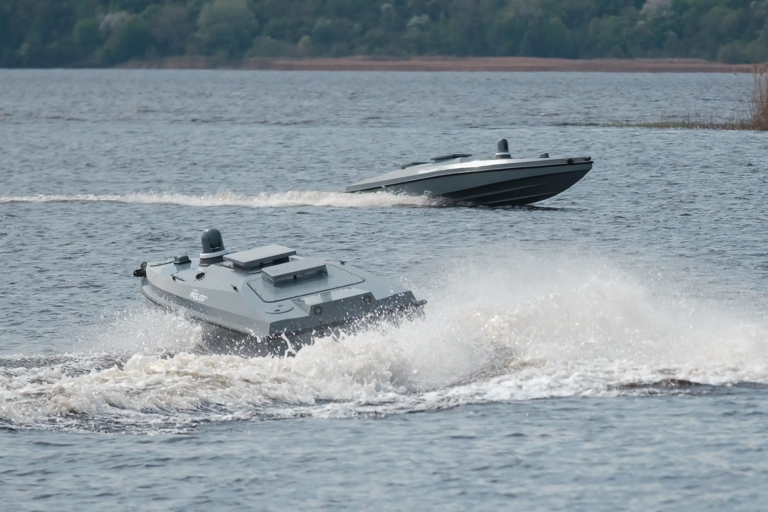 Two grey drone boats sail in water with a line of trees seen in the background.