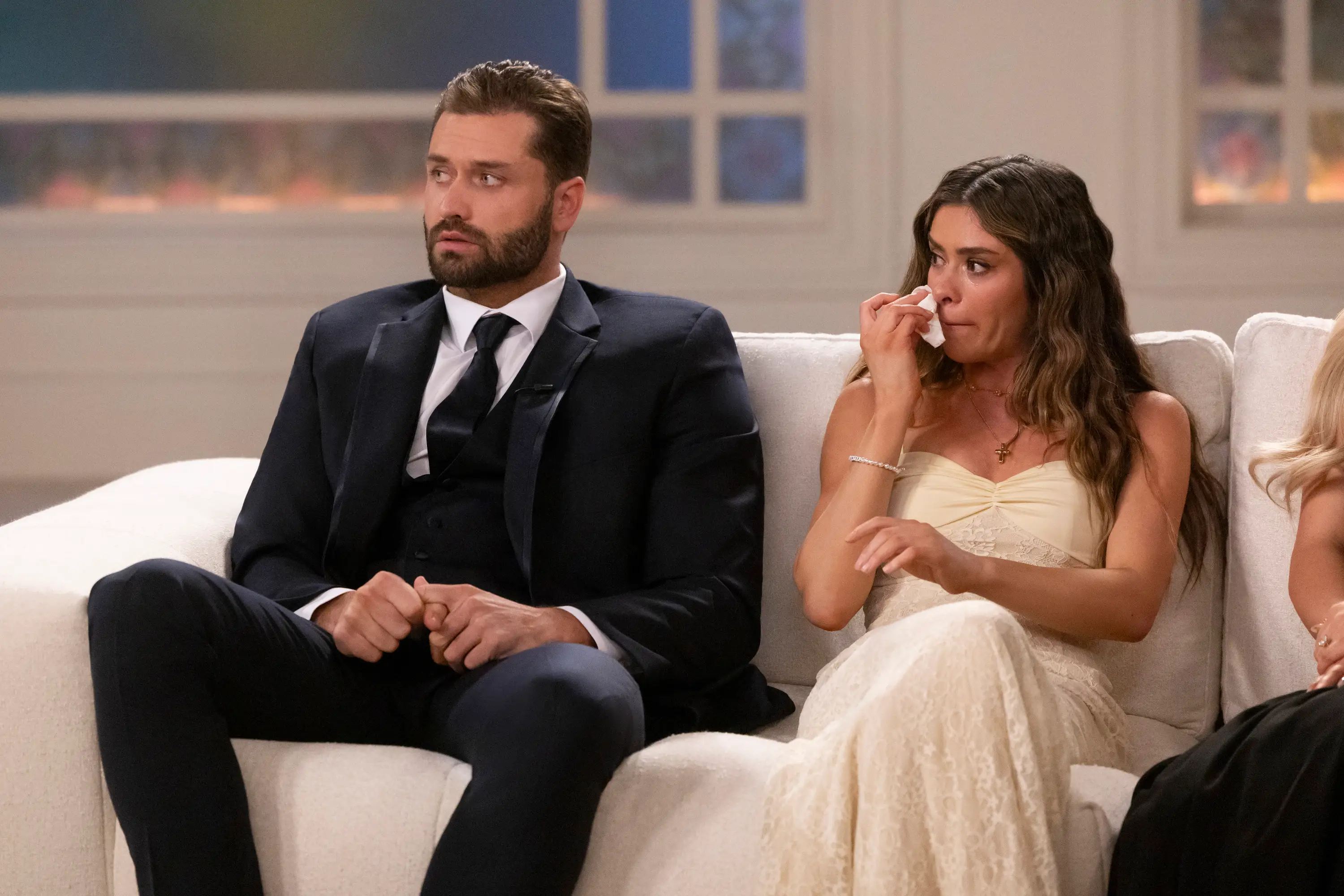 A man in a suit sits on a couch next to a woman in a white dress who's wiping away tears.