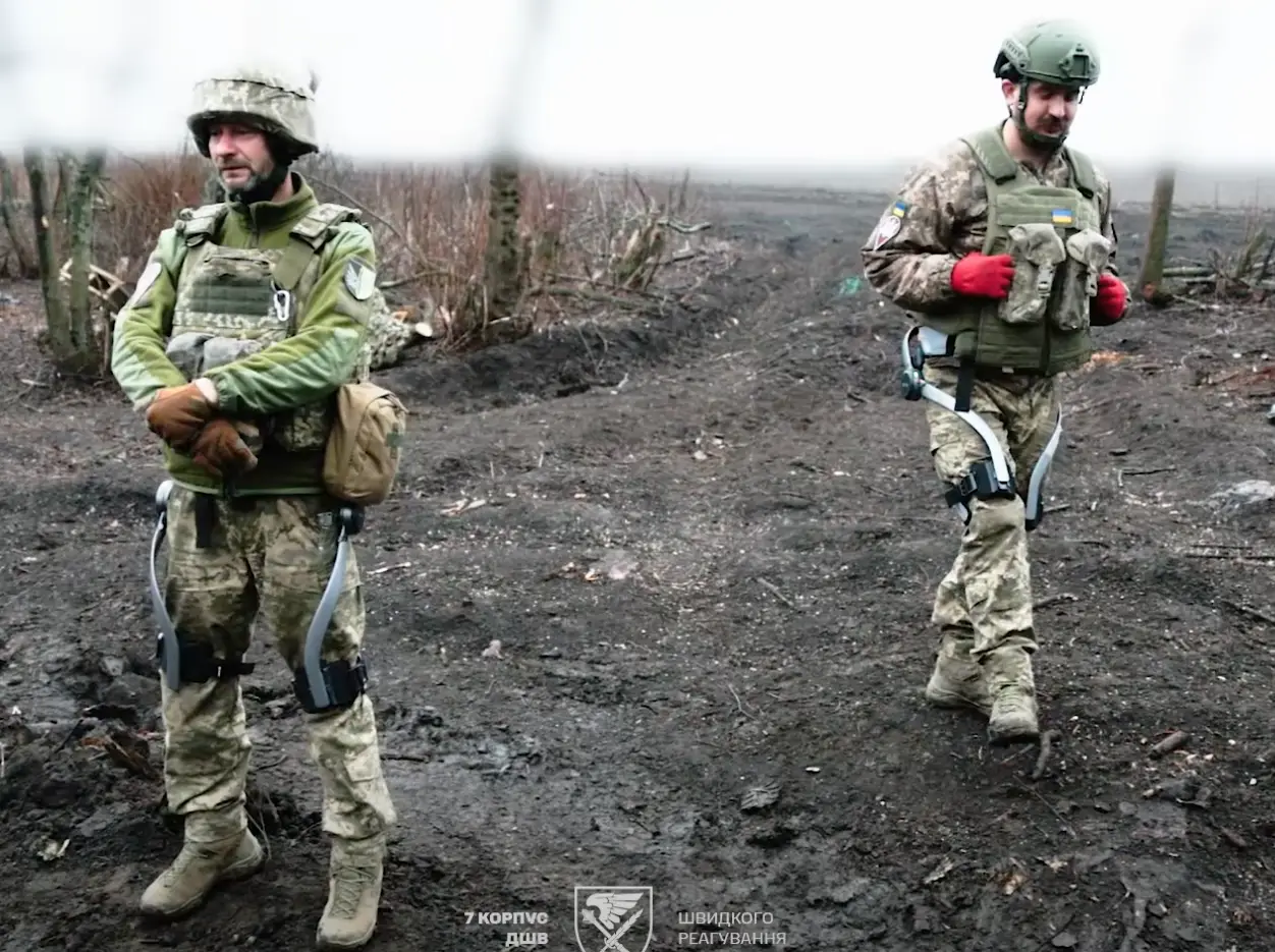 Two soldiers in military fatigues, tactical vests, and exoskeletons walk the battlefield.