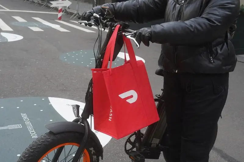 A red DoorDash bag hangs from the handlebars of a e-bike as someone wearing black pants, a black coat, and black gloves pushes the bike.
