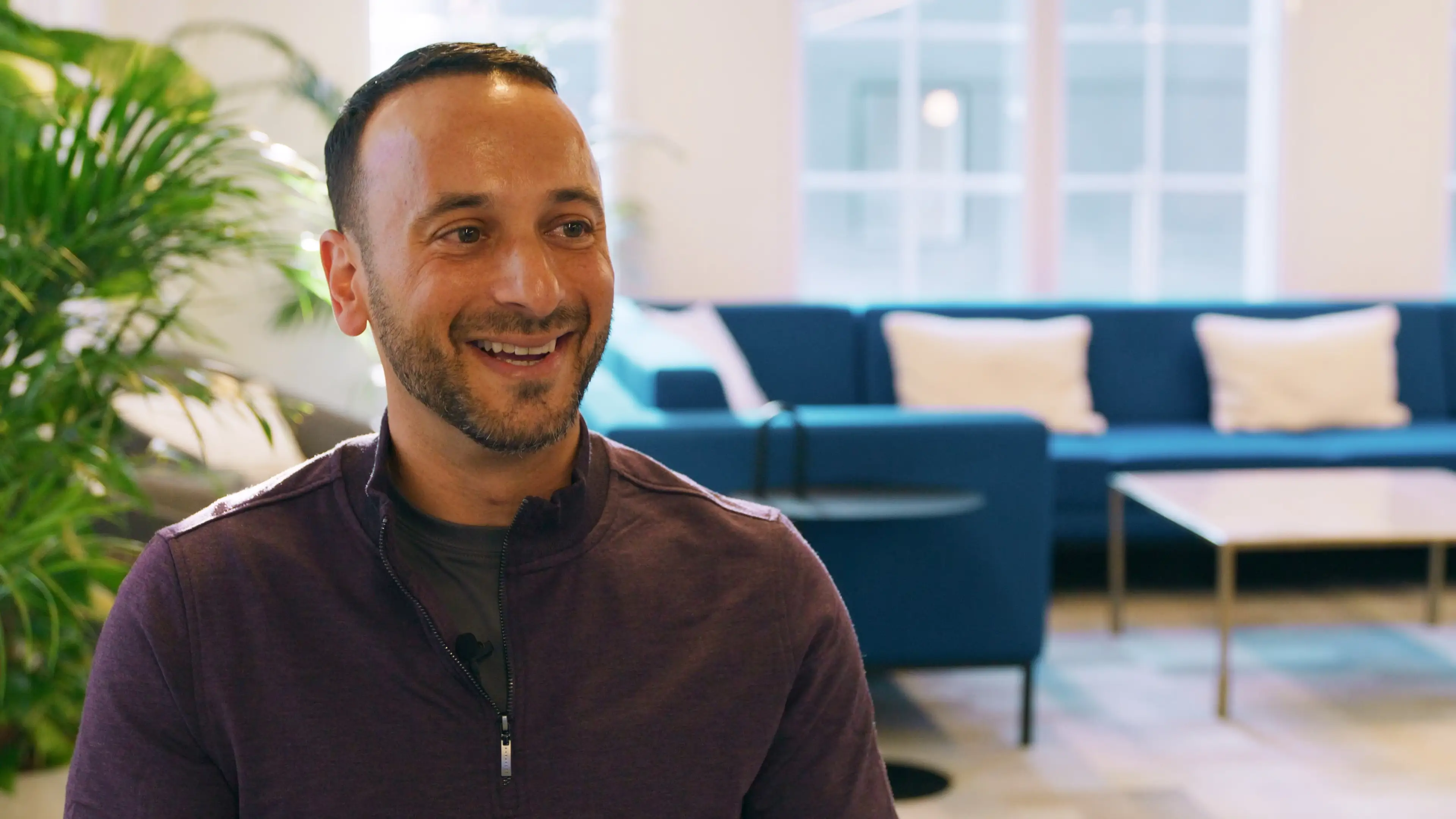 A smiling man sits in a bright office lounge with blue couches and a large leafy plant in the background.