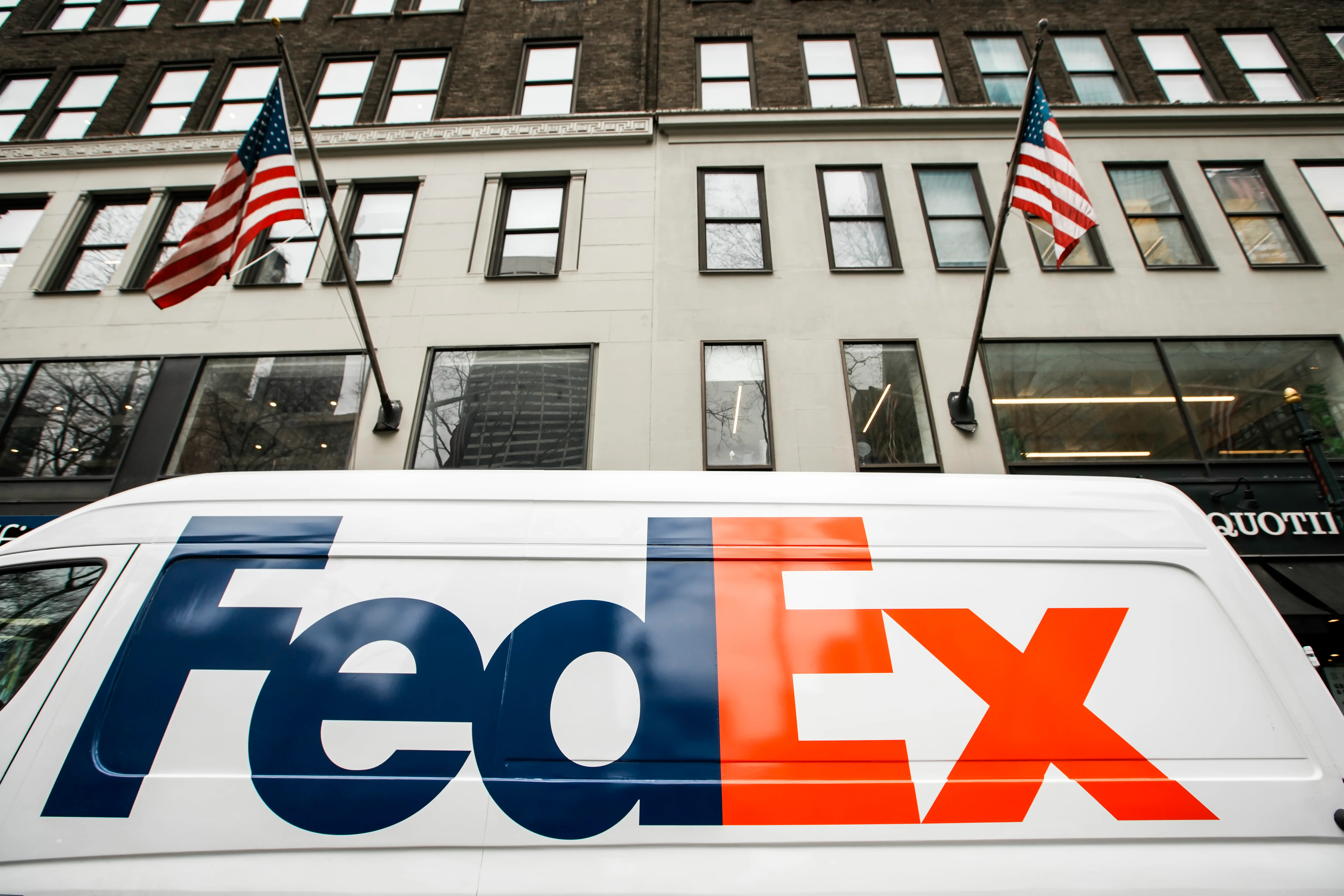 A FedEx van sits below two American flags outside of a building.
