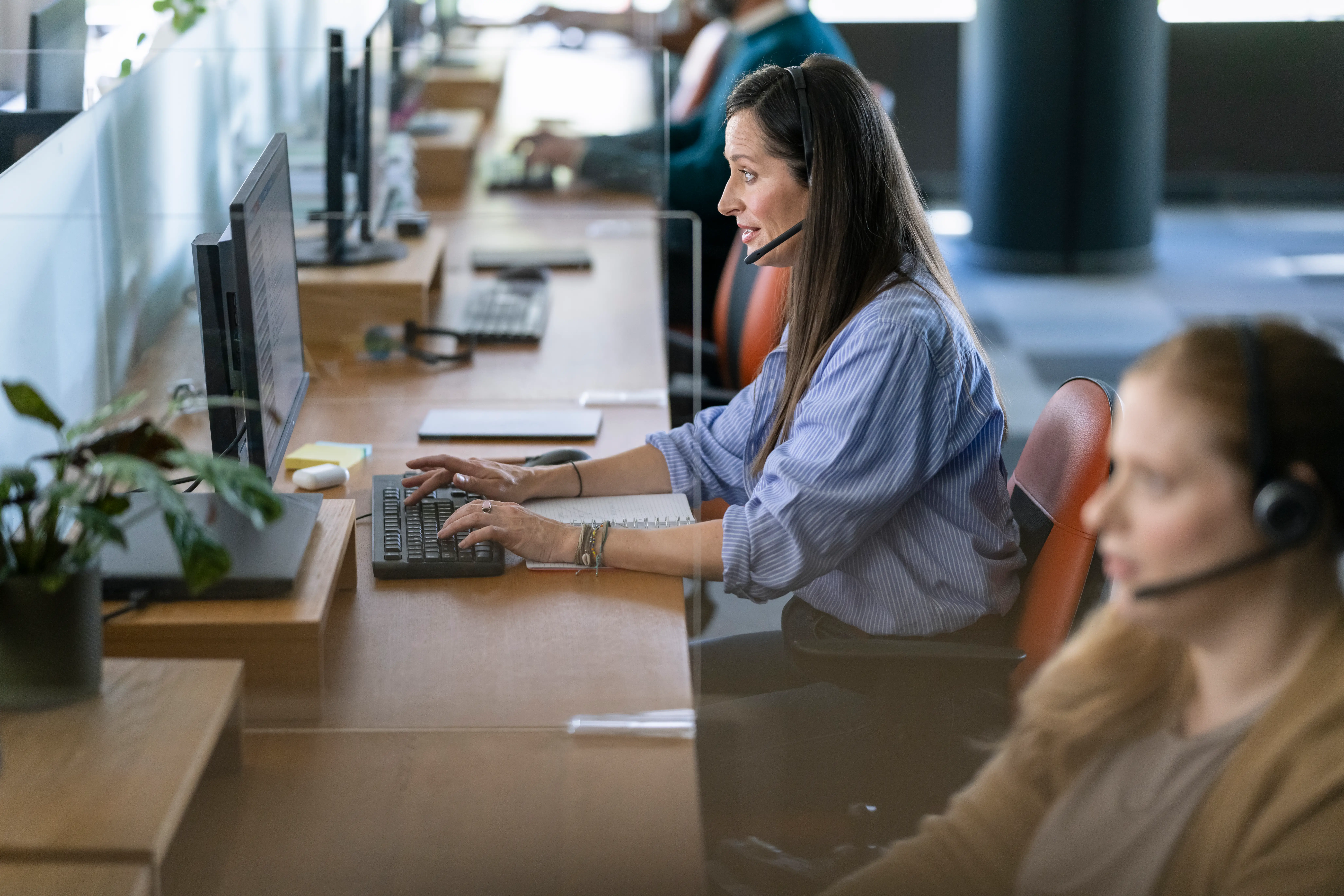 A woman in a blue blouse sits at her computer with a headset on for sales.