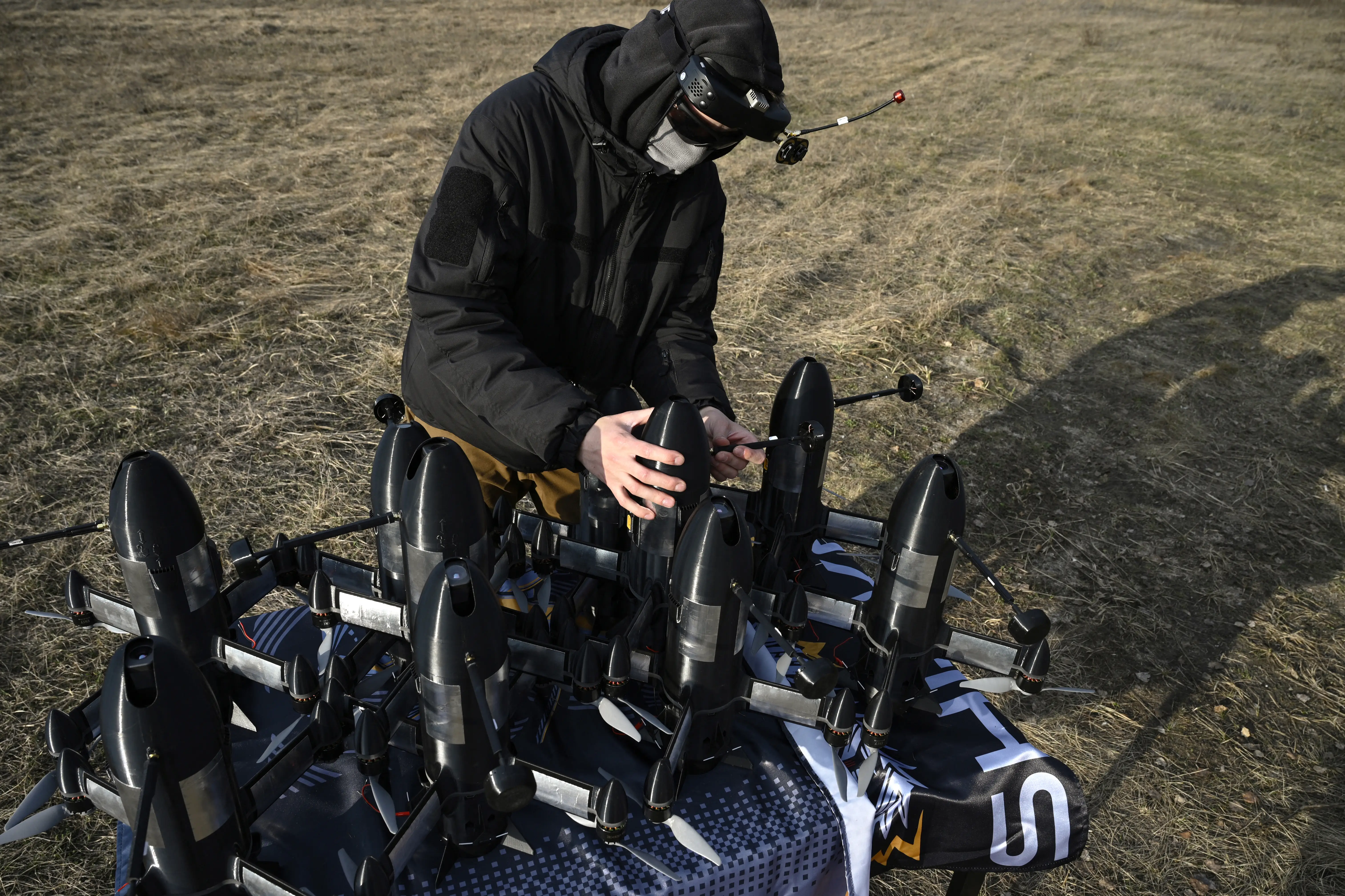 A man in a black outfit kneels on the grass, holding on to one of a host of small black interceptor drones that are on the ground