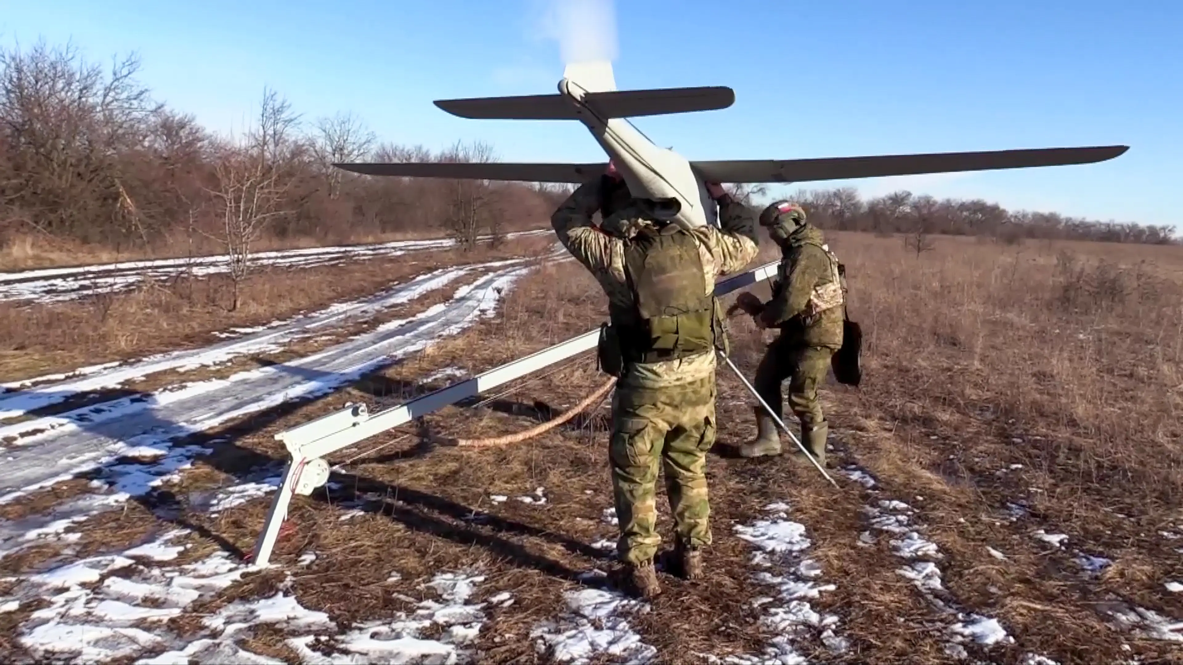 Two men in camouflage gear in a snowy brown field under a blue sky with one holding a large grey drone