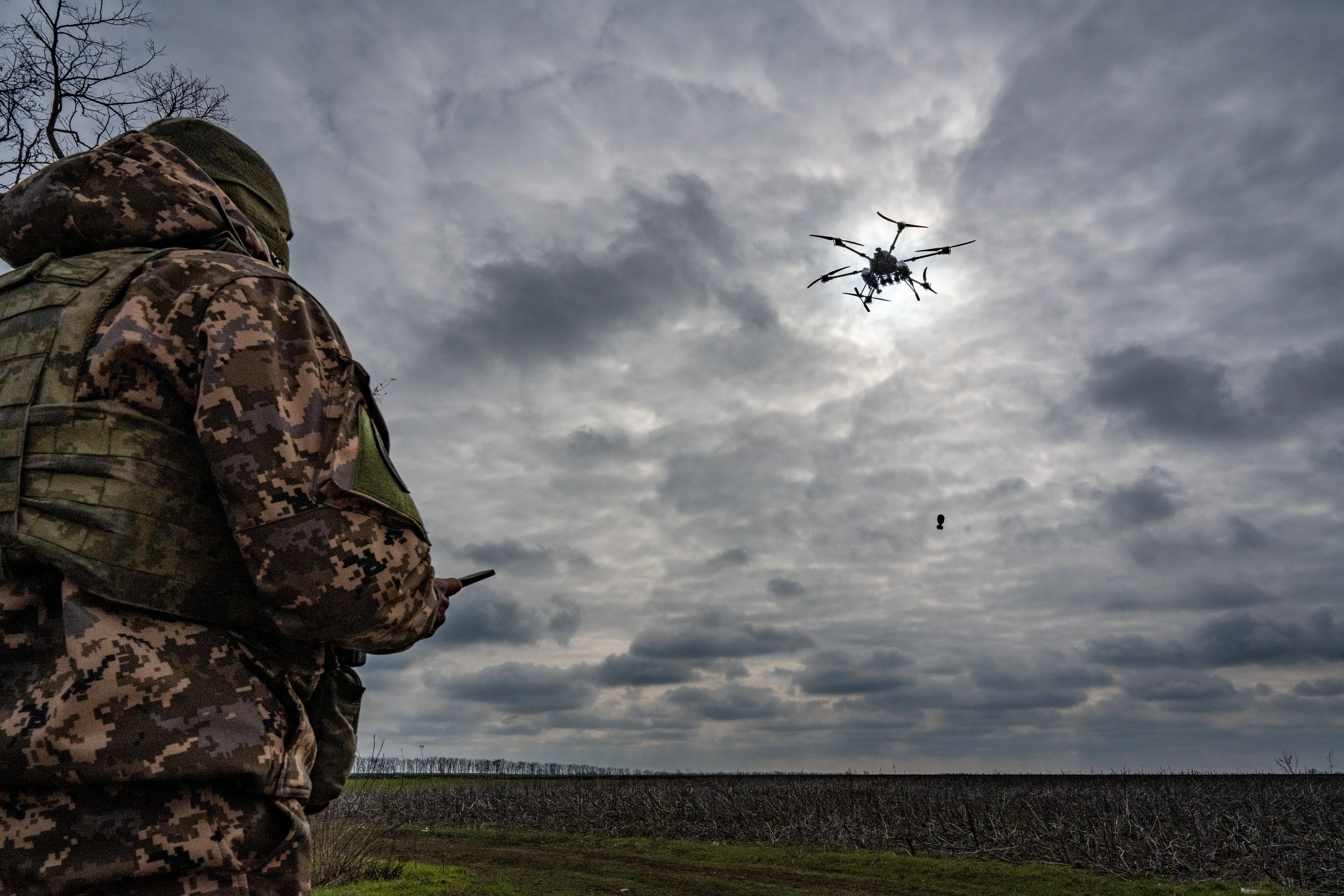 A man in camouflage stands with his back turned looking at a field and cloudy sky with a drone in it
