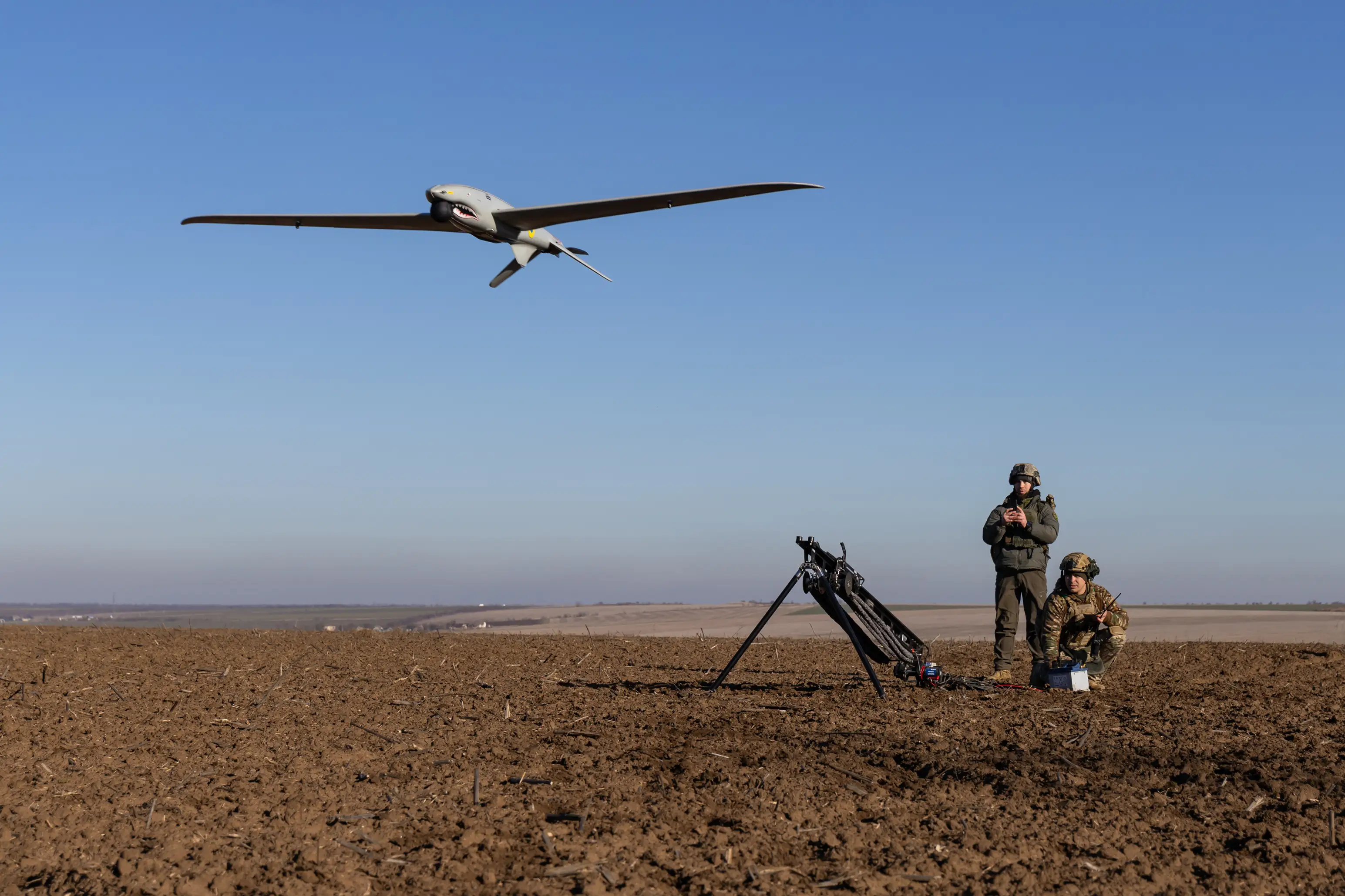 Two figures in green look at a grey drone flying in the air, over a brown field and under a blue sky