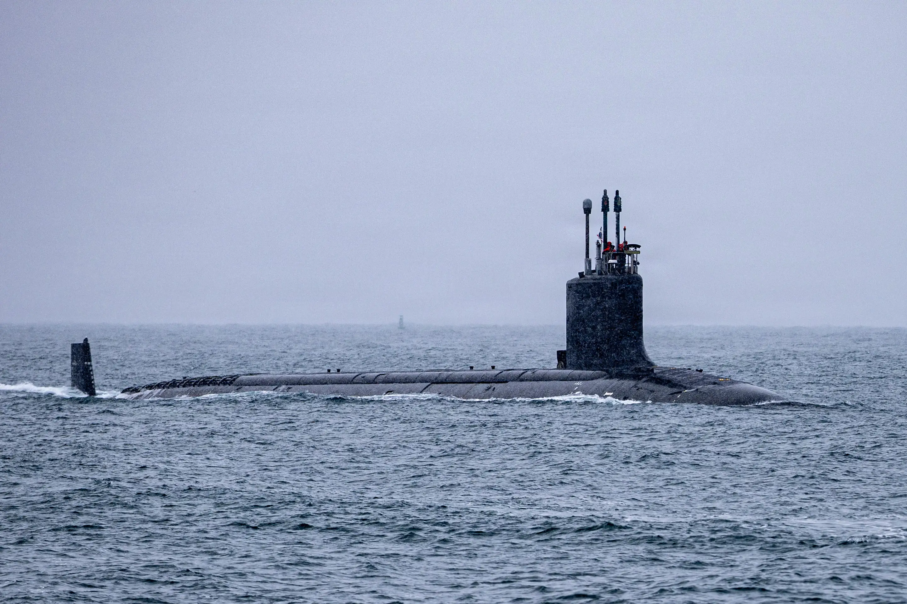 A long black submarine sails in dark blue water with a cloudy sky in the background.