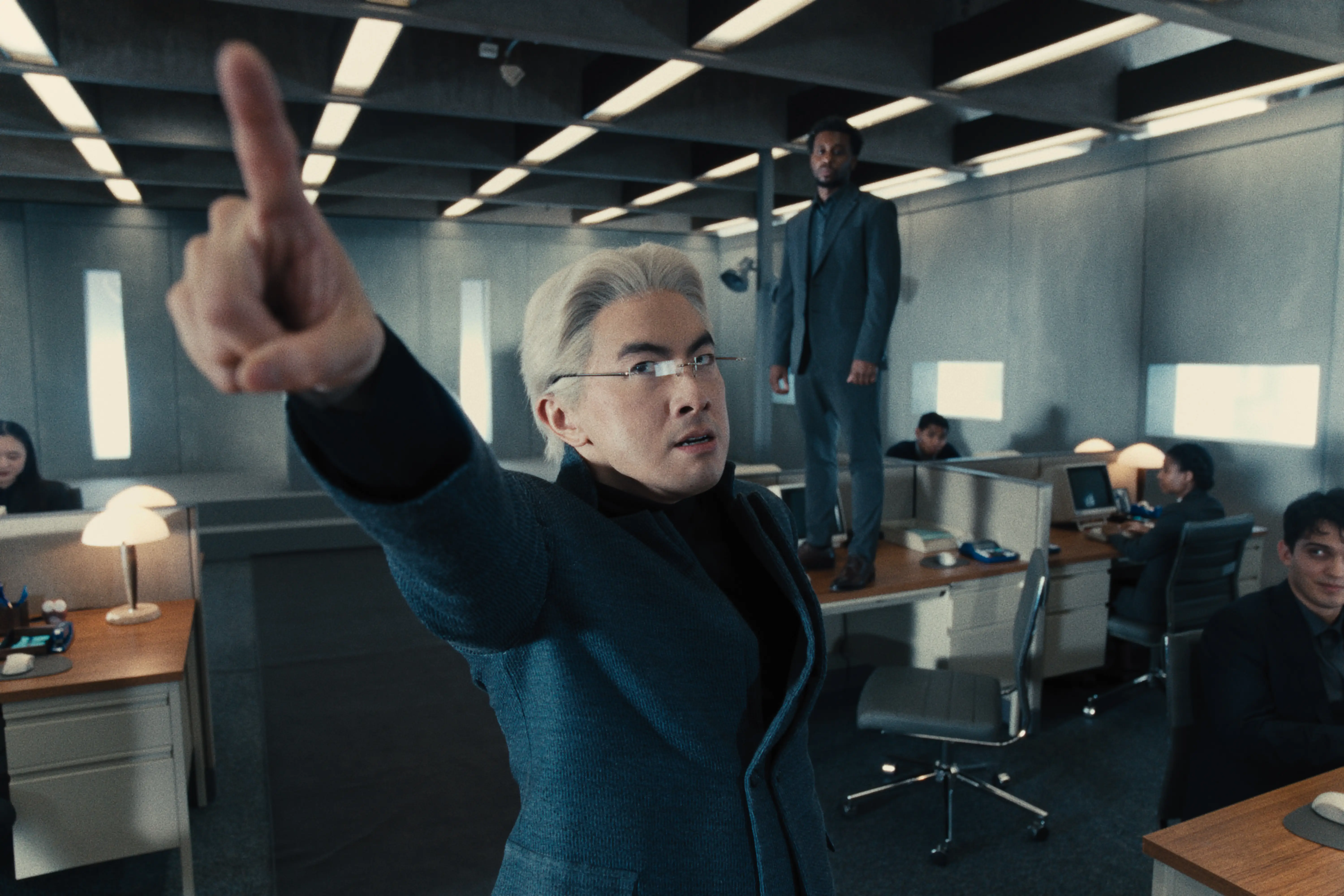 Man in suit standing on desk with another man in a suit in front of him pointing in the air.